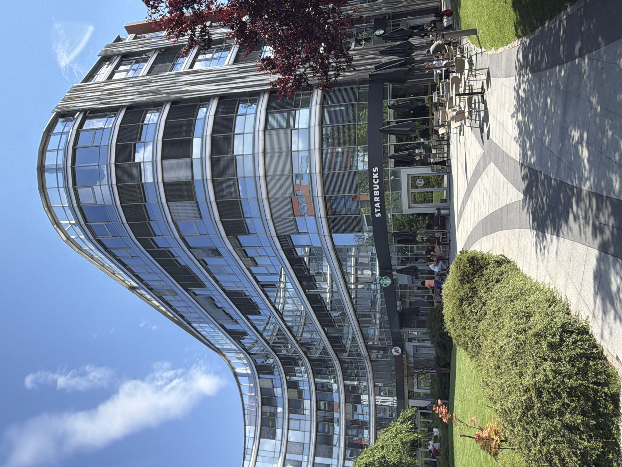 The image depicts an exterior view of a modern, curved glass-fronted building with a Starbucks on the ground floor. Several people are visible seated at tables outside the Starbucks, suggesting a casual social gathering or break. The time of day appears to be daytime, with bright sunlight casting shadows on the paved walkway. The weather seems to be clear and sunny, with a few wispy clouds in the blue sky. A lush green lawn and manicured bushes are present, adding to the pleasant outdoor setting. The building's architecture is characterized by its flowing, curved facade and numerous windows that reflect the sky and surrounding environment. The Starbucks signage is clearly visible. The context provided indicates the location is Sofia, Bulgaria. The overall impression is a lively urban scene on a pleasant day.