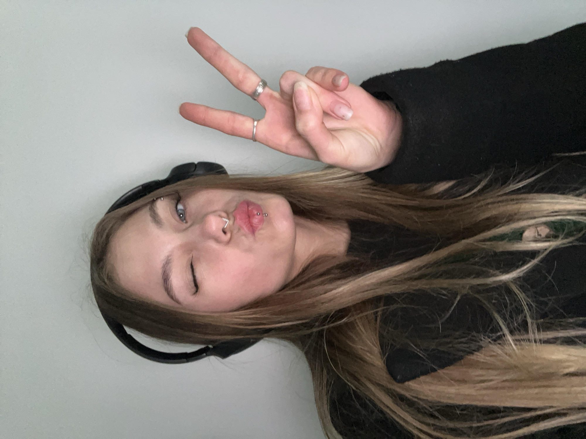 A young woman is positioned with her face and body turned to the left, and she is holding up a hand in a peace sign. She has blonde hair, is wearing headphones, and is winking with her right eye. She has piercings in her eyebrow, nose, and lips, and rings on her fingers. The background is a plain, light-colored wall. There is no information about Фоки, Russia, present in the image.