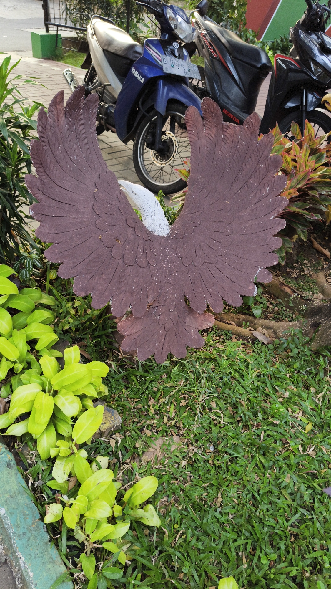 An eye-level, medium shot captures a statue of a brown eagle with its wings spread, positioned in front of two parked motorcycles, a Yamaha and a Honda. The eagle statue is made of what appears to be concrete and is located on a patch of green grass. To the left of the eagle, bright green foliage is visible. In the background, the motorcycles are parked on a paved area. The Yamaha is blue and white, with the license plate "B 6474 FUM". The Honda is black with red accents. The scene appears to be outdoors, likely in a public space within the city of South Jakarta, Indonesia, possibly a park or a residential complex's common area.