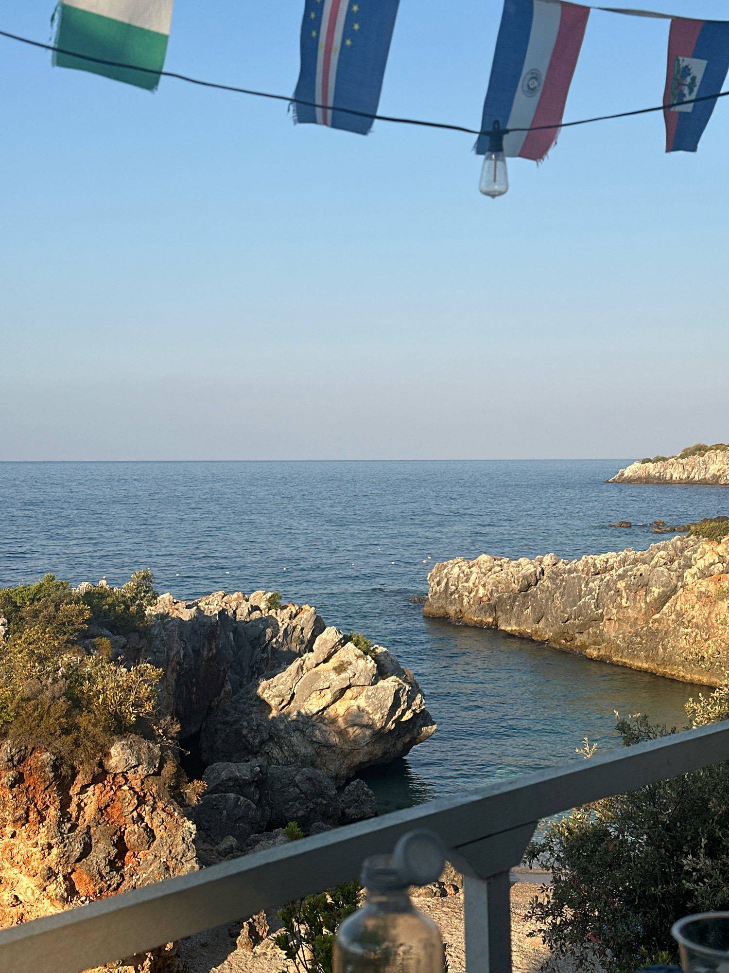 The image shows a body of water with rocky shores, seen from a vantage point with a railing in the foreground. Above the water, a string of small flags and a lightbulb are visible against a clear blue sky. The flags appear to be of different nations, and the overall scene suggests an outdoor setting, possibly a cafe or restaurant overlooking the sea. The water is calm and reflects the blue of the sky. The rocks are irregular in shape and covered with sparse vegetation. The lightbulb is a vintage style bulb.