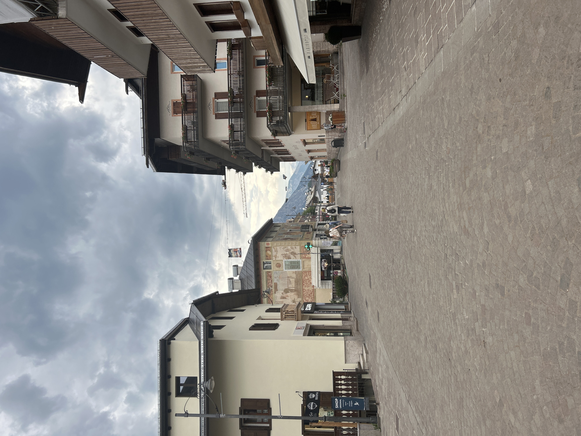 This image captures a street scene in what appears to be a European town, possibly in the Alps, given the mountainous backdrop visible in the distance. The perspective is from street level, looking down a cobblestone street lined with multi-story buildings. The architecture suggests a traditional style, with wooden balconies, pitched roofs, and some buildings featuring decorative murals.
Several people are visible walking along the street, engaged in casual activity. The street itself is paved with cobblestones, some arranged in a patterned curve. Shops and restaurants are interspersed along the street level of the buildings, indicated by signage and display windows. One sign reads "MARINOLDI". Another sign has "BOTTEGA" followed by what looks like "VENETA". Below that, "SCARPA" is visible, and further down, "REGINA" and "SHOP".
The sky is overcast with heavy, gray clouds, suggesting a potentially damp or cool day. The time of day is difficult to determine precisely due to the clouds, but it appears to be daytime, possibly late afternoon or early evening given the lighting. The overall mood is calm and leisurely, reflecting a typical day in a quaint town. The presence of a construction crane in the background hints at ongoing development or renovation in the area. The scene is static, focusing on the architecture and the ambiance of the street.