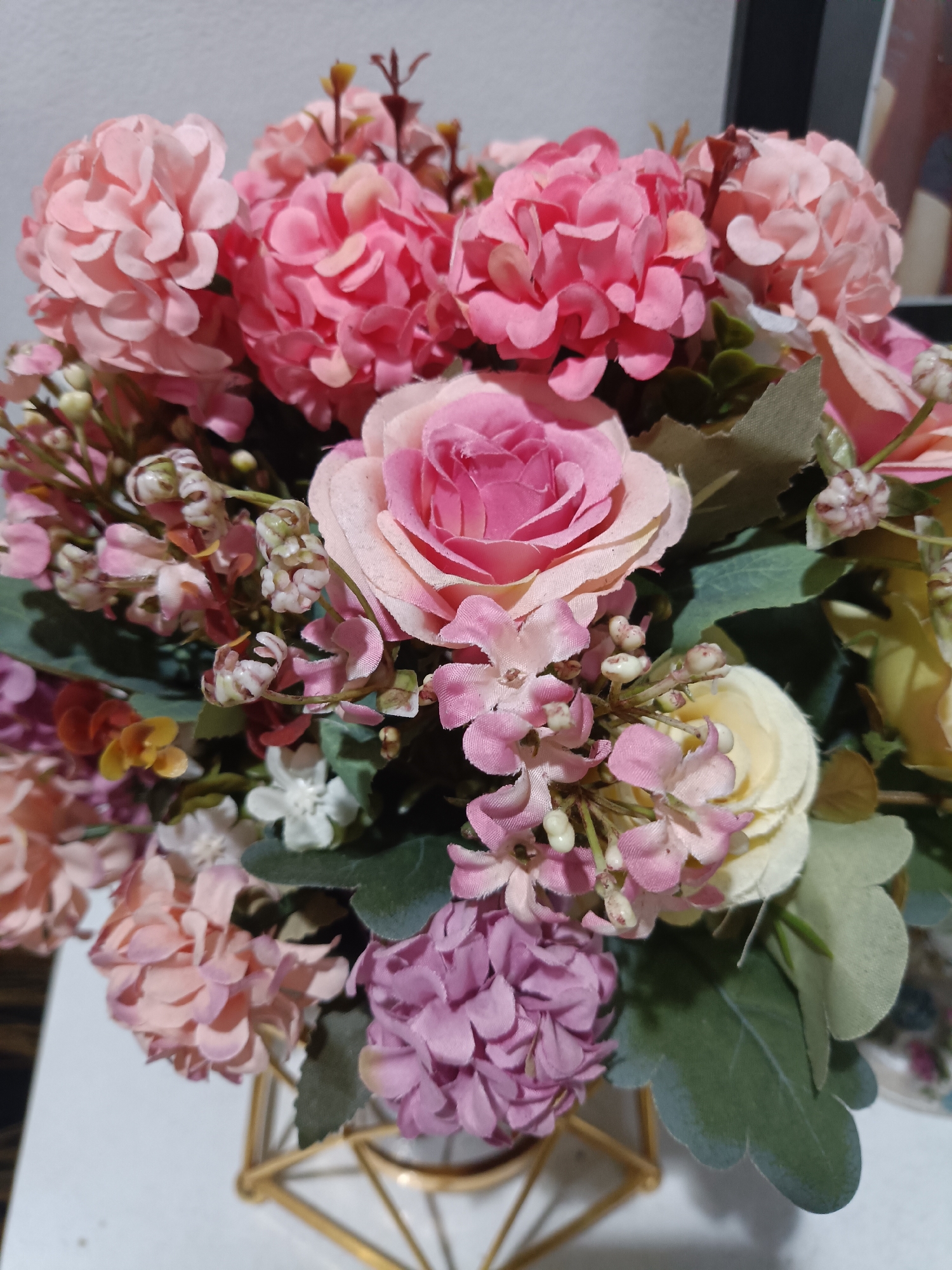 This is a close-up, vertically oriented, eye-level shot of a bouquet of artificial flowers. The flowers are predominantly in shades of pink, with some lighter pink, cream, and purple hues mixed in. There's a large, open pink rose in the center, surrounded by smaller, clustered flowers that resemble hydrangeas and smaller spray-like blossoms with tiny white buds. Green artificial leaves are interspersed throughout the arrangement. The bouquet is partially visible inside a gold, geometric-shaped vase. The background is a plain, light-colored wall, and a sliver of what appears to be a framed picture or artwork is visible in the upper right corner, suggesting an indoor setting. The lighting is soft and even, indicating it is likely daytime, possibly with ambient indoor lighting. No people are visible, and no specific event or activity is taking place. The overall impression is one of decorative arrangement, likely for home or event decor. The location context of West Jakarta, Indonesia, does not provide any readily discernible visual cues within the image itself.