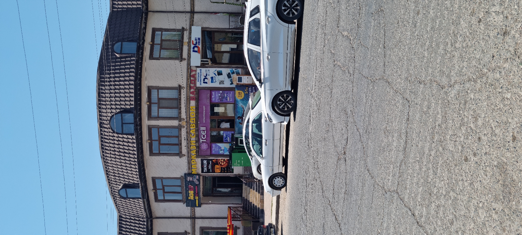 The image shows a street scene in the city of Ismoili Somoni, Tajikistan. In the foreground, two white cars are parked on a cracked asphalt road. Behind the cars is a two-story building with a dark tiled roof and dormer windows. The building's facade is light-colored and features several windows on both levels. Below the upper windows, there are multiple storefronts with signage. One storefront has a sign that reads "MARKAZI SAVDOI BAXT" and another displays the TCELL logo. Adjacent to these, there is a store with a "DC CITY" sign and a promotional display with images of electronic devices. To the left, a store labeled "TANUR" is visible, with a menu or advertisement displayed outside. Stairs lead up to one of the entrances of the building. The sky is clear and blue.