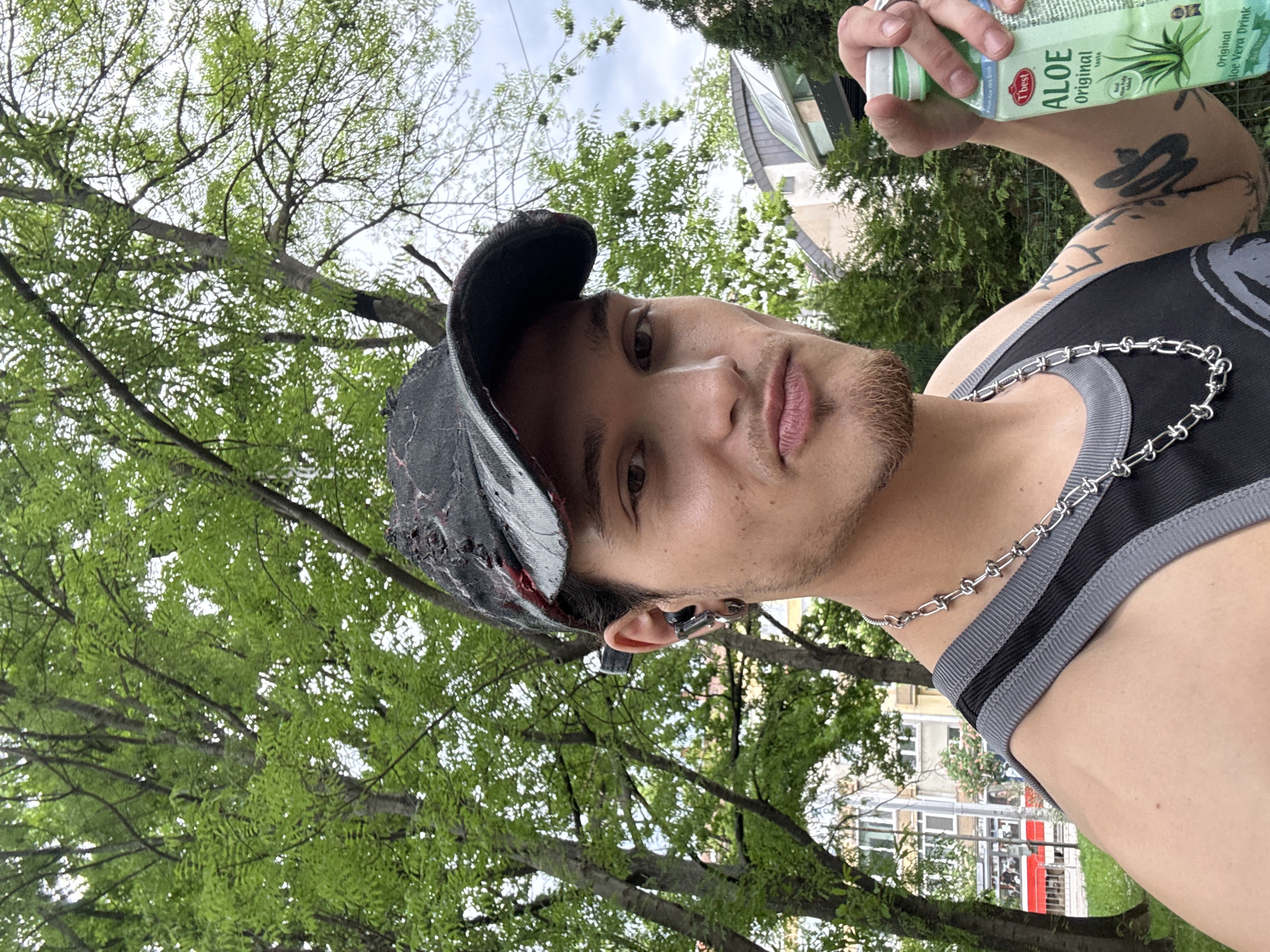 A selfie shot from a low angle captures a young adult male outdoors. The man is wearing a distressed black baseball cap, a black tank top with grey trim, and a silver chain necklace. Tattoos are visible on his right arm. He holds a bottle of "The Best Aloe Original Aloe Vera Drink" in his right hand. He has short dark hair, light brown skin, and stubble. He is wearing an earbud in his left ear. The background consists of green trees with leaves, a building, and a patch of grass behind him. The presence of a streetcar and architectural style suggest a location within Sofia, Bulgaria.