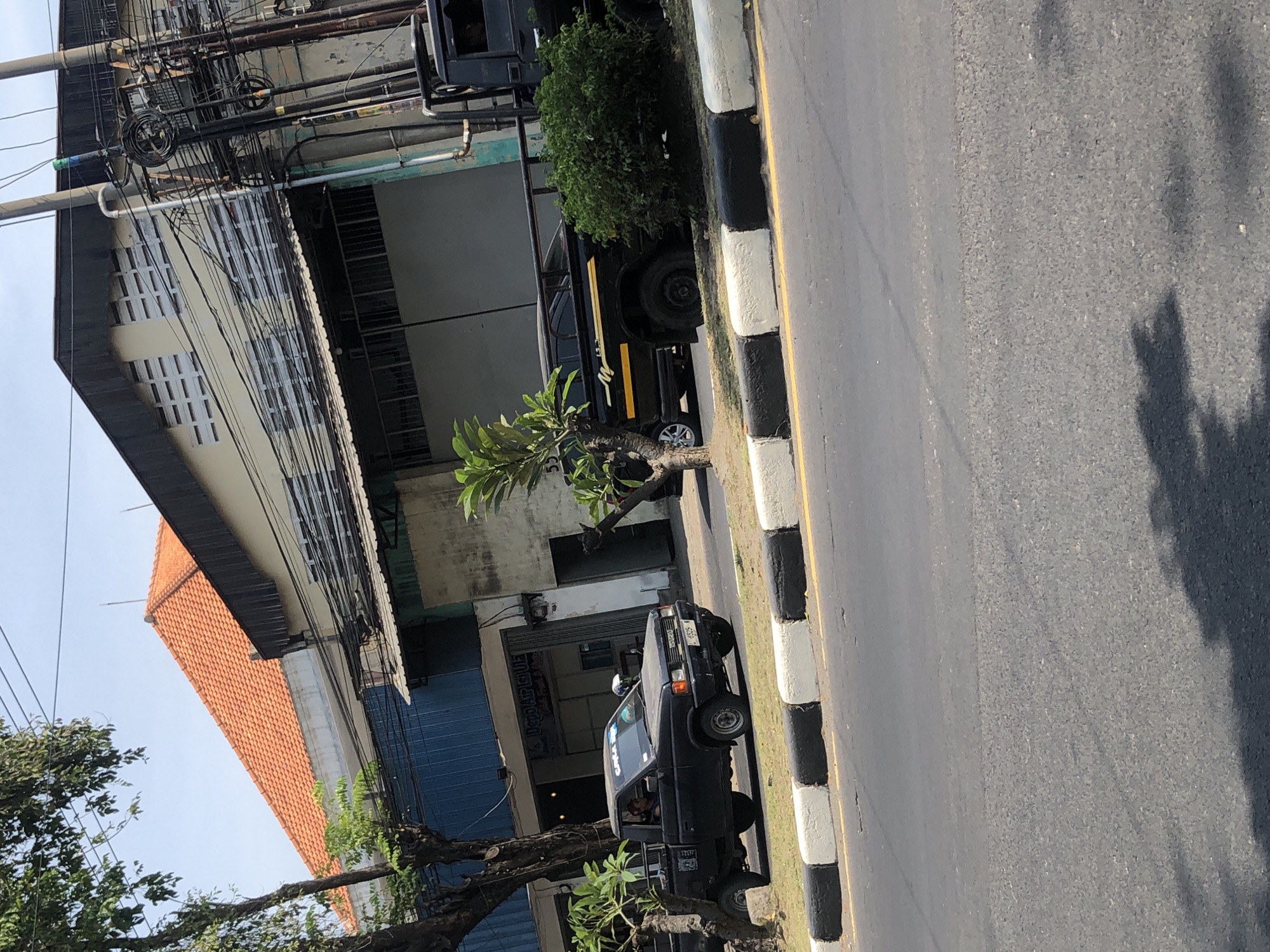 The image depicts a street scene in Larangan, Indonesia, on a sunny day. In the foreground, a black pickup truck with a driver is parked on the side of the road, next to a curb with alternating black and white painted sections. The truck has a license plate that reads "B 1325 WFN". Further down the road, another black truck, possibly a larger utility vehicle with a metal railing structure on its bed, is visible. The background shows a building with an orange tiled roof and a light-colored facade, interspersed with blue corrugated metal siding. Electrical wires and poles are prominent along the side of the building, indicating urban infrastructure. Trees with green foliage are present on the left and right sides of the frame, with one tree in the foreground casting a shadow on the road. The scene appears to be a commercial or industrial area given the building style and the presence of multiple vehicles.