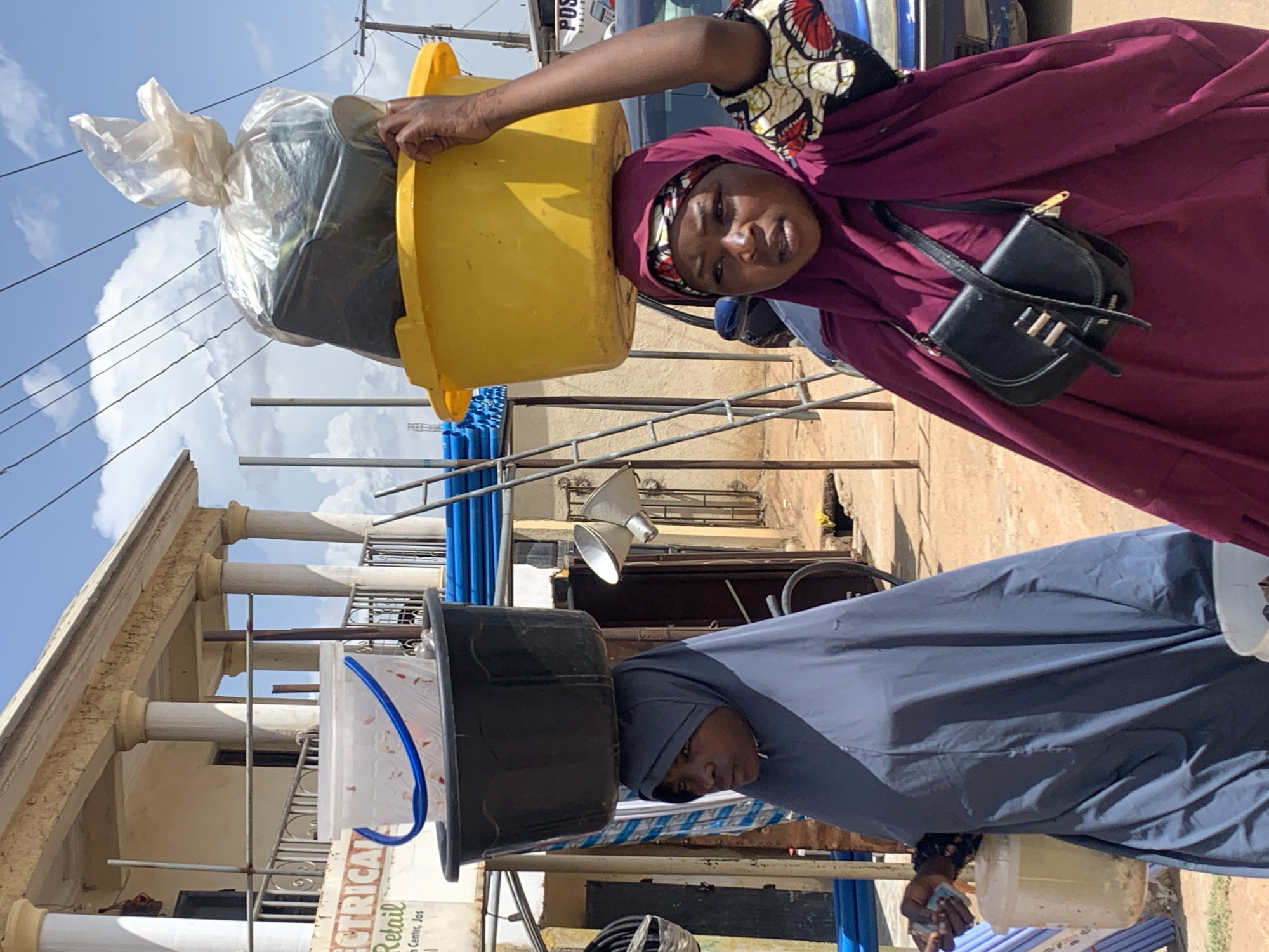 The image, set in Gura Top, Nigeria, captures a sunlit street scene featuring two women transporting items on their heads. The sky is clear with scattered white clouds, indicating bright daylight.
In the foreground on the right, a woman in a deep maroon headscarf and matching long dress looks directly at the viewer. She balances a large yellow basin on her head, which contains a clear plastic bag filled with what appears to be merchandise or personal belongings. Her left hand steadies the basin, and a small black shoulder bag with gold accents hangs by her side. She wears a patterned top underneath her outer garment.
Further back on the left, another woman, dressed in a greyish-blue headscarf and attire, walks balancing a black basin on her head. Inside this black basin is a smaller, clear plastic container with a blue handle. She also holds a white container in her right hand. Both women are engaged in the common practice of carrying goods on their heads, likely as part of daily errands or market activities.
The background features a two-story building with distinct white columns and light-colored walls, possibly a commercial establishment or residence. Scaffolding is visible around parts of the building. Blue pipes or barrels are stacked horizontally on the ground. Power lines and utility poles crisscross the upper part of the frame. A blue vehicle, partially visible behind the first woman, has "POS" written on its side. On the lower left, a partial sign reads "RICAL," "Retail," and "Jos," with "Inc" also visible, suggesting an "Electrical Retail" business located in Jos, Nigeria.