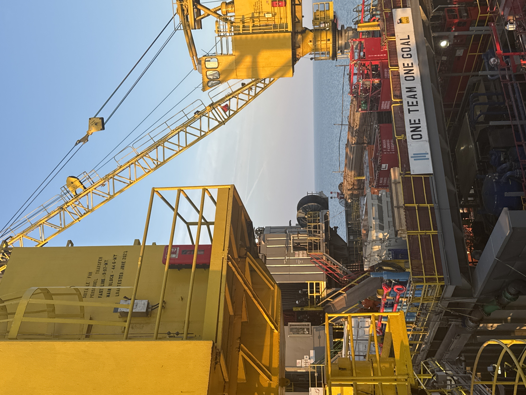 The image shows an offshore oil rig with yellow cranes and structures against a clear blue sky and the ocean. Several workers in hard hats and safety vests are visible on the platforms and walkways. There are also various industrial equipment, pipes, and containers on the rig. A banner with the text "ONE TEAM ONE GOAL" is prominently displayed. The lighting suggests it is daytime, likely late afternoon given the warm hue of the sunlight. The scene appears to be an operational industrial environment at sea.