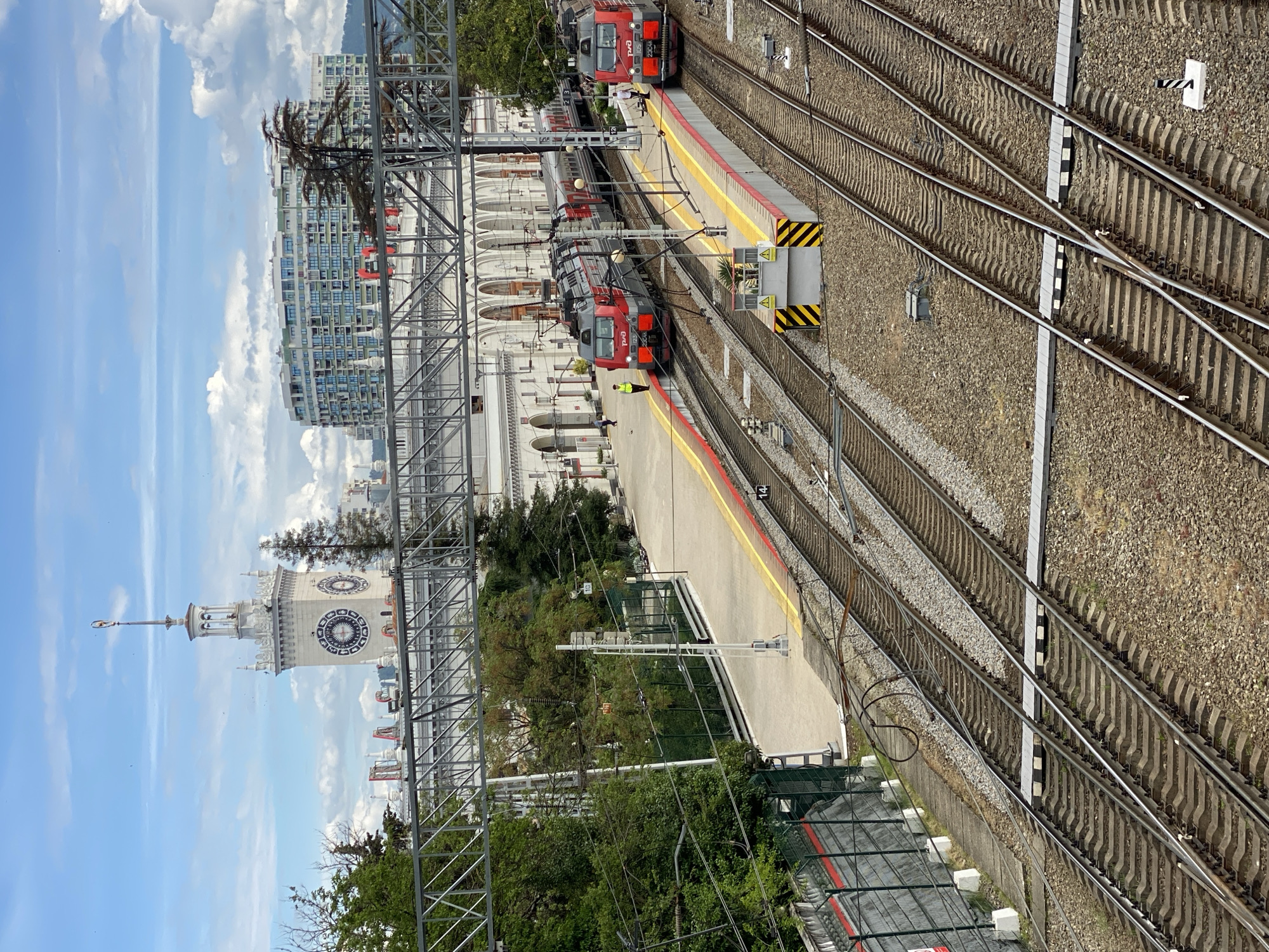The image depicts the Sochi Railway Station in Sochi, Russia. Two red electric trains are visible at the platforms. A few people are present on the platform near the train. The railway tracks converge and run alongside the platforms. The station building, a large white structure with arched windows, is visible in the background. A clock tower is also prominent. The sky is partly cloudy, with blue sky visible. Trees and greenery are present around the station area. Overhead electrical cables and metal structures are visible above the tracks and platform.