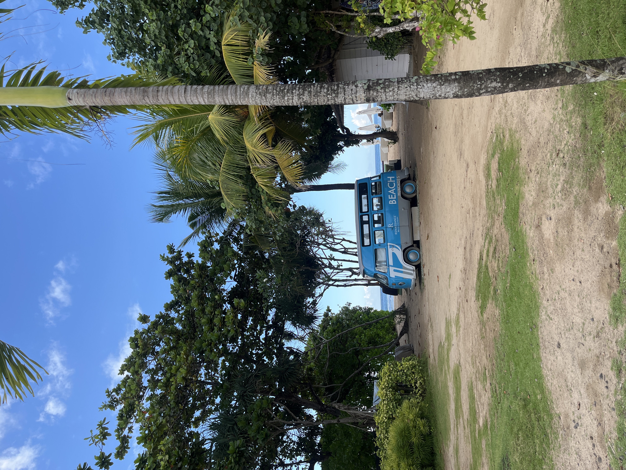 The image shows a beach scene in Kuta Selatan, Indonesia. A blue 'Beach Munchies' food truck is parked on a sandy path lined with trees, leading towards a beach with white umbrellas visible in the distance. The sky is clear blue with scattered clouds. A tall palm tree is prominent in the foreground.