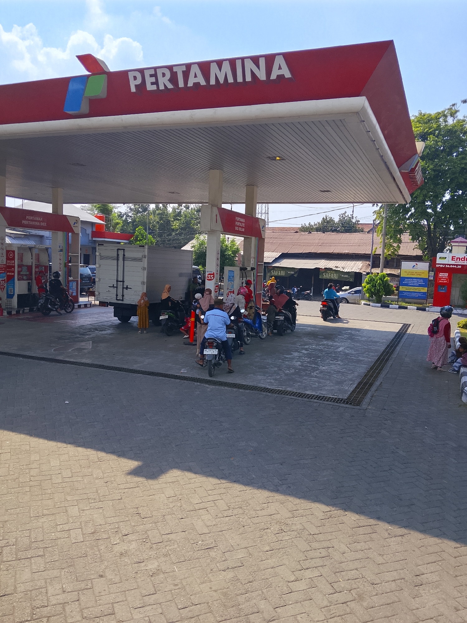 The image depicts a sunny day at a Pertamina gas station, likely located in Kudu, Indonesia. The main structure is a red canopy with "PERTAMINA" written in large white letters. Beneath the canopy, several fuel pumps are visible. A white truck with a box trailer is parked at one of the pumps. Multiple people are present, some on motorcycles and others standing. A few individuals are wearing headscarves. The ground is paved with brick-like stones. In the background, there are trees, buildings with corrugated metal roofs, and a bright blue sky with scattered clouds. One sign in the background reads "Enduro Motor Oil". Another sign displays "Pertamax Pertalite Bio Solar". Several people appear to be waiting to refuel their motorcycles. The overall scene suggests a busy but orderly refueling operation on a typical day.