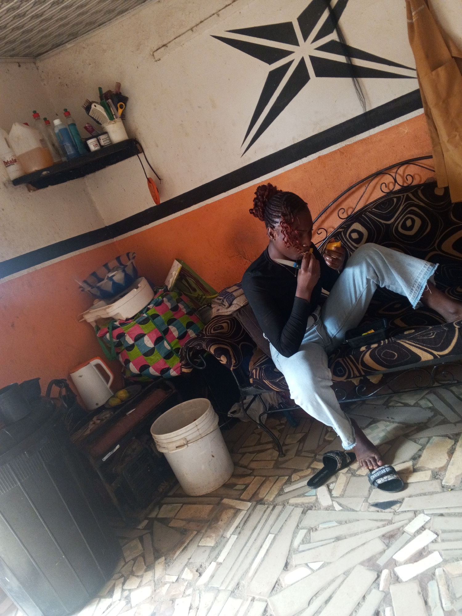 The scene appears to be the interior of a room in Gwong, Nigeria. A young woman with braided hair sits on a patterned couch, wearing a black top and light blue jeans. She is holding a piece of orange fruit and appears to be eating it. A container, possibly containing the rest of the fruit, is visible on the couch near her.
The room has a shelf with various containers on the wall and a large black and white geometric design. There is a white bucket and other miscellaneous items, including a bag, a kettle, and what appears to be a generator, on the floor. The floor is tiled with irregular shapes.