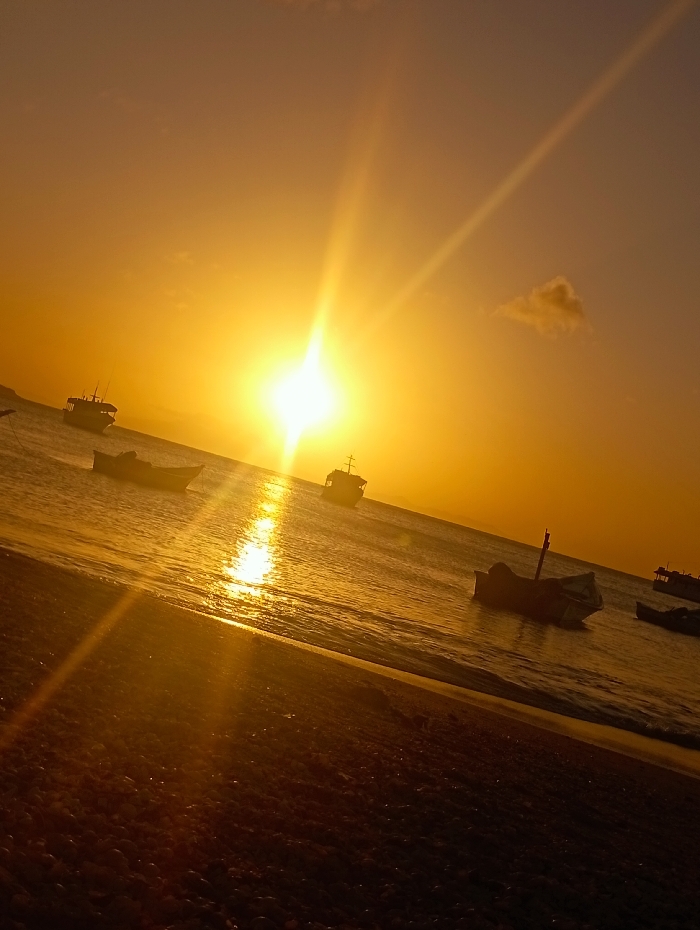 Golden sunset illuminates a beach scene. Several boats are anchored in the calm waters near the shoreline. The sun, a bright focal point, casts long rays across the water and the sand. A single, small cloud floats in the upper right. The beach is textured with sand and small stones. The image is bathed in warm, golden light.