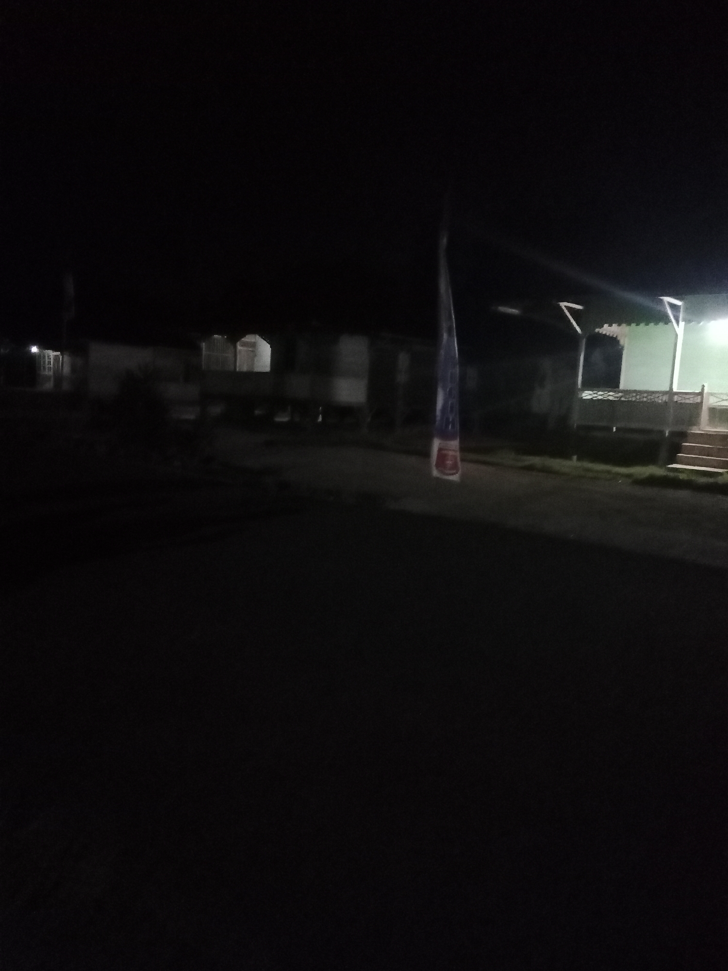 The image, taken at night in Liwa, Indonesia, captures a dimly lit street scene with several buildings visible. In the foreground, a dark road stretches into the distance. To the left, a portion of a building with illuminated windows can be seen. In the center, a tall, vertical banner with red and blue markings hangs, illuminated by an unseen light source. To the right, a building with a green roof and an open-air structure supported by pillars is partially visible, with stairs leading up to it. The overall atmosphere is dark and quiet, suggesting a residential or semi-commercial area at rest.