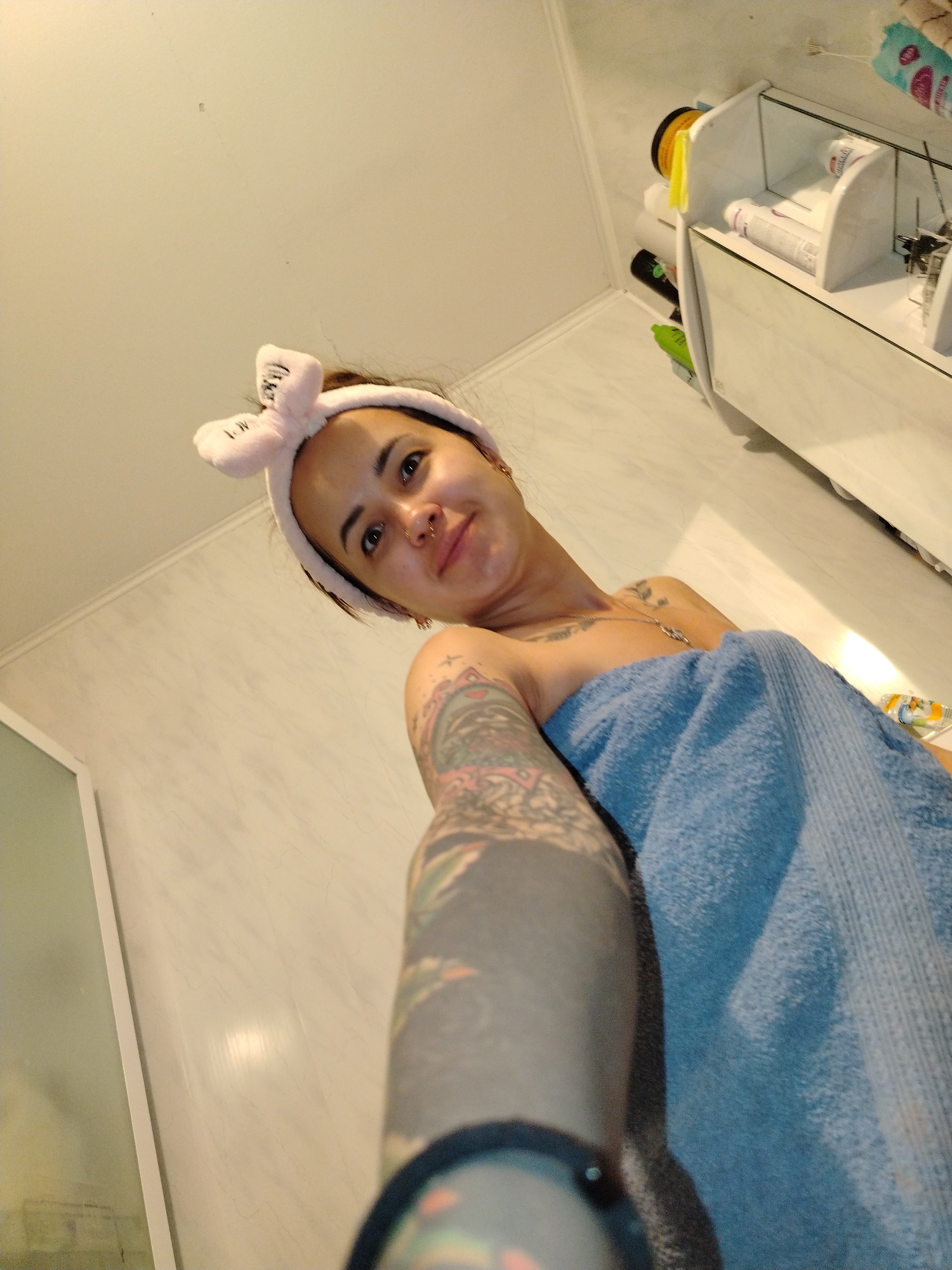 The image depicts a young woman wearing a pink bow headband and a blue towel. She has tattoos on her arm and chest. The woman is looking up at the camera, which is positioned below her, suggesting a selfie taken from a low angle. The background appears to be a bathroom with tiled walls and shelves containing various toiletries. There is a reflection of the woman and the bathroom environment in a mirror. The overall impression is that the woman is in a bathroom, possibly after a shower. There is no information in the image that can be used to determine the location in the city of Noviy, Russia.