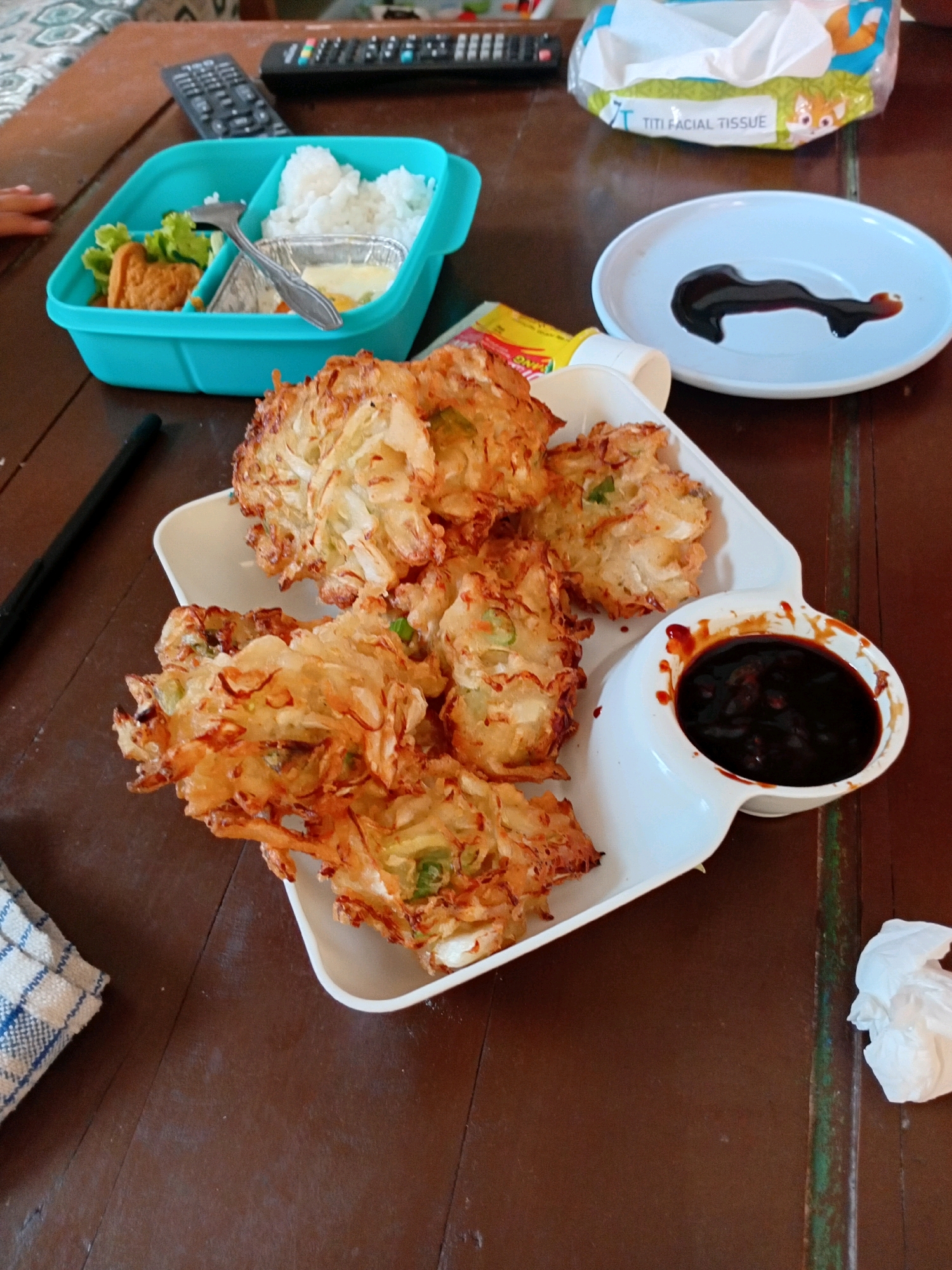 This is a photograph of a meal on a wooden table, likely taken in Gebang, Indonesia. In the foreground, a white platter holds several pieces of golden-brown fried food, possibly bakwan or vegetable fritters, served with a small white bowl filled with dark brown sauce. To the left of the fritters is a turquoise lunchbox divided into compartments. One compartment contains white rice, another holds a piece of fried chicken and lettuce, and a third contains a small foil container with a sauce or dip. A silver fork rests in the lunchbox. In the background, two TV remote controls are visible, along with a package of TITI facial tissues featuring a cartoon fox. A white crumpled tissue is also present on the table. The lighting suggests an indoor setting, possibly a home dining area. The overall scene depicts a casual meal.