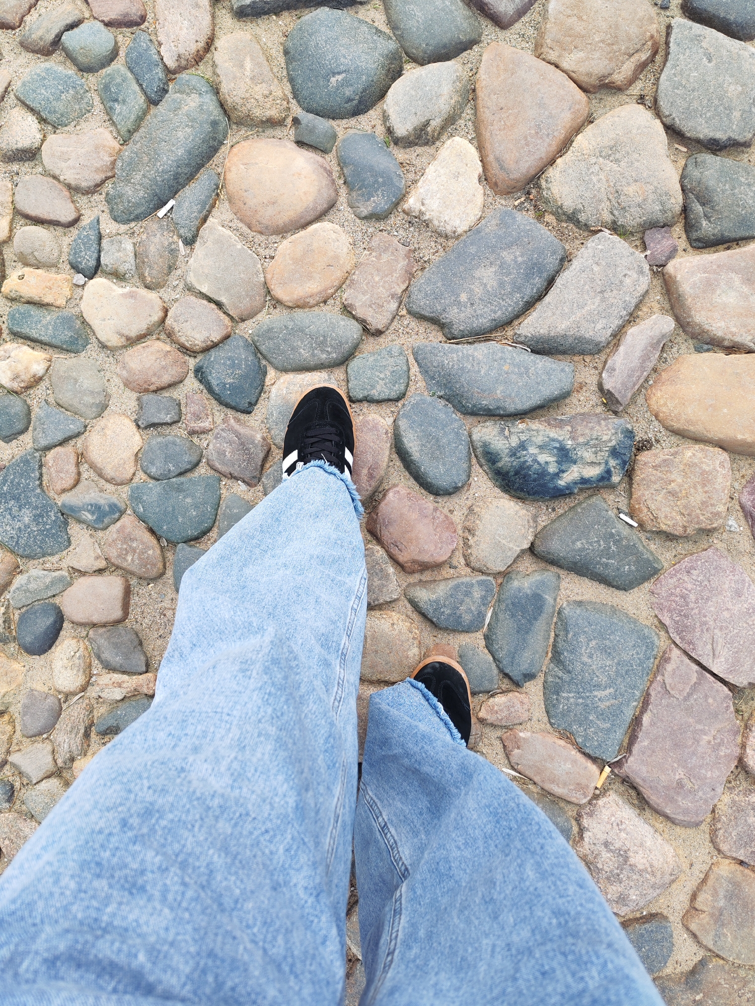 A pair of light blue denim jeans and black sneakers with white stripes are worn by an unseen person walking on a cobbled path. The cobblestones are of various sizes and colors, ranging from gray and brown to pink and tan, and are interspersed with sand. The scene appears to be outdoors, possibly in the city of Bol'shie Mkhi, Russia, given the cobbled pathway and the natural elements. The perspective is from above, looking down at the person's legs and feet as they walk. The lighting suggests it is daytime, with natural light illuminating the scene.