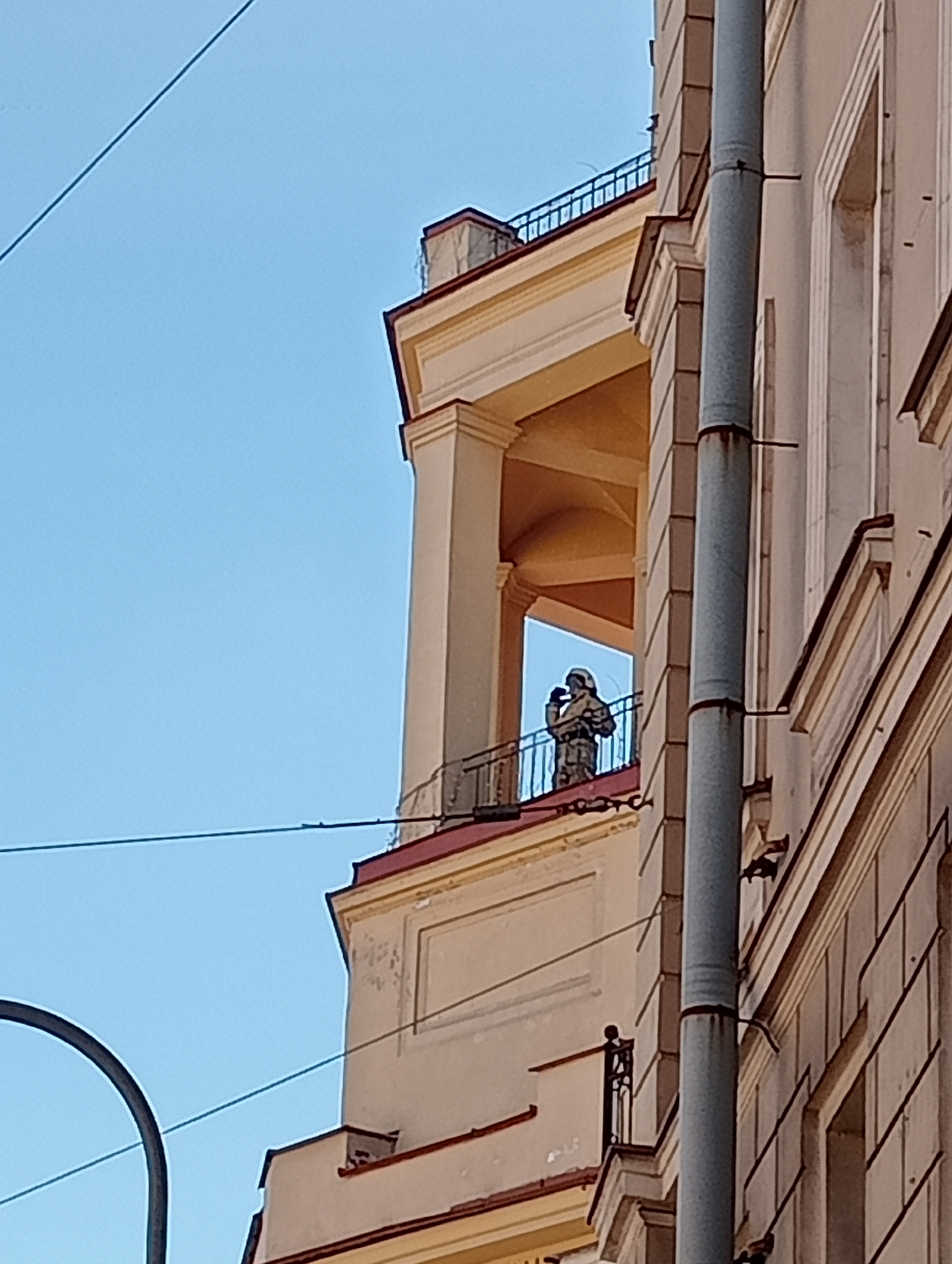 A person in camouflage clothing, holding a rifle, stands on a balcony of a cream-colored building with classical architectural details. The building has a large grey pipe running vertically down its facade. The sky is a clear, light blue. This scene is located in Saint Petersburg, Russia, near Nevsky Prospect, possibly in the vicinity of Anichkov Bridge, known for its historical architecture.