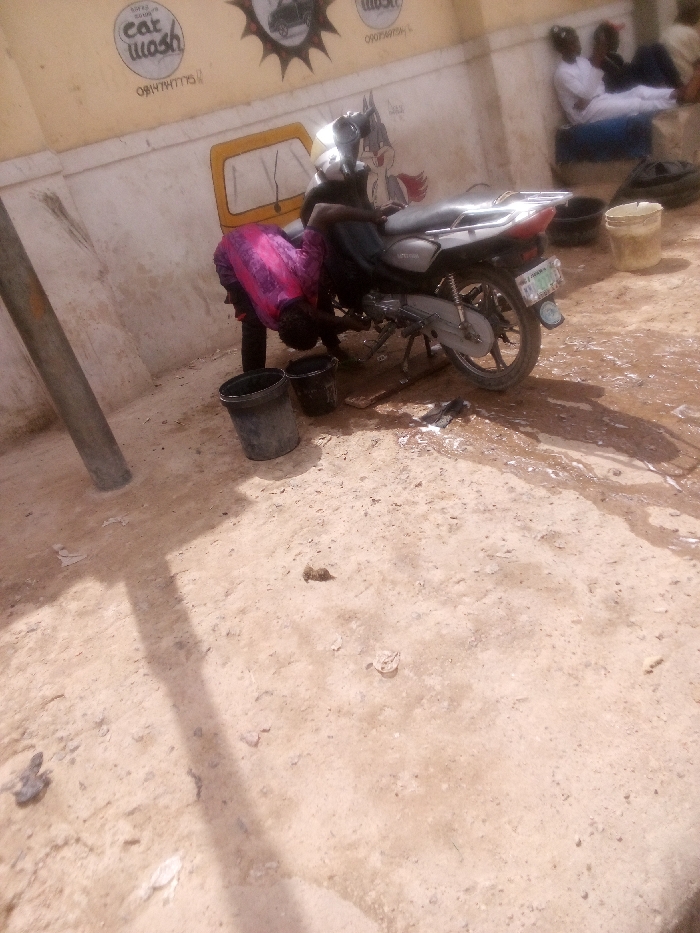 The image displays an outdoor scene in Tudun Murtala, Nigeria, depicting a motorcycle wash operation. In the foreground, the ground consists of light-colored, dry earth, interspersed with wet patches and soapy runoff originating from the washing activity. A grey, cylindrical pole is vertically positioned on the left side.
A single individual, dressed in a bright pink patterned top over dark trousers, is actively washing a silver-grey motorcycle positioned on its kickstand. This person is bent over, focused on cleaning the lower engine and chassis components of the vehicle. Two dark-colored plastic buckets are present on the ground beside the motorcycle, one containing water or soap solution. A dark rag or sponge lies on the wet ground near the motorcycle's rear wheel, which bears a visible Nigerian license plate.
In the background, a light yellow building wall serves as the backdrop. It is decorated with painted graffiti, including a circular "Car Wash" logo, several phone numbers (one clearly visible as "08147747775"), a cartoon character resembling Bugs Bunny, and a yellow outline of a vehicle. To the right, three additional individuals are observed seated or reclining against the wall; two are attired in light-colored, possibly white, traditional garments. They appear to be observing the washing activity.