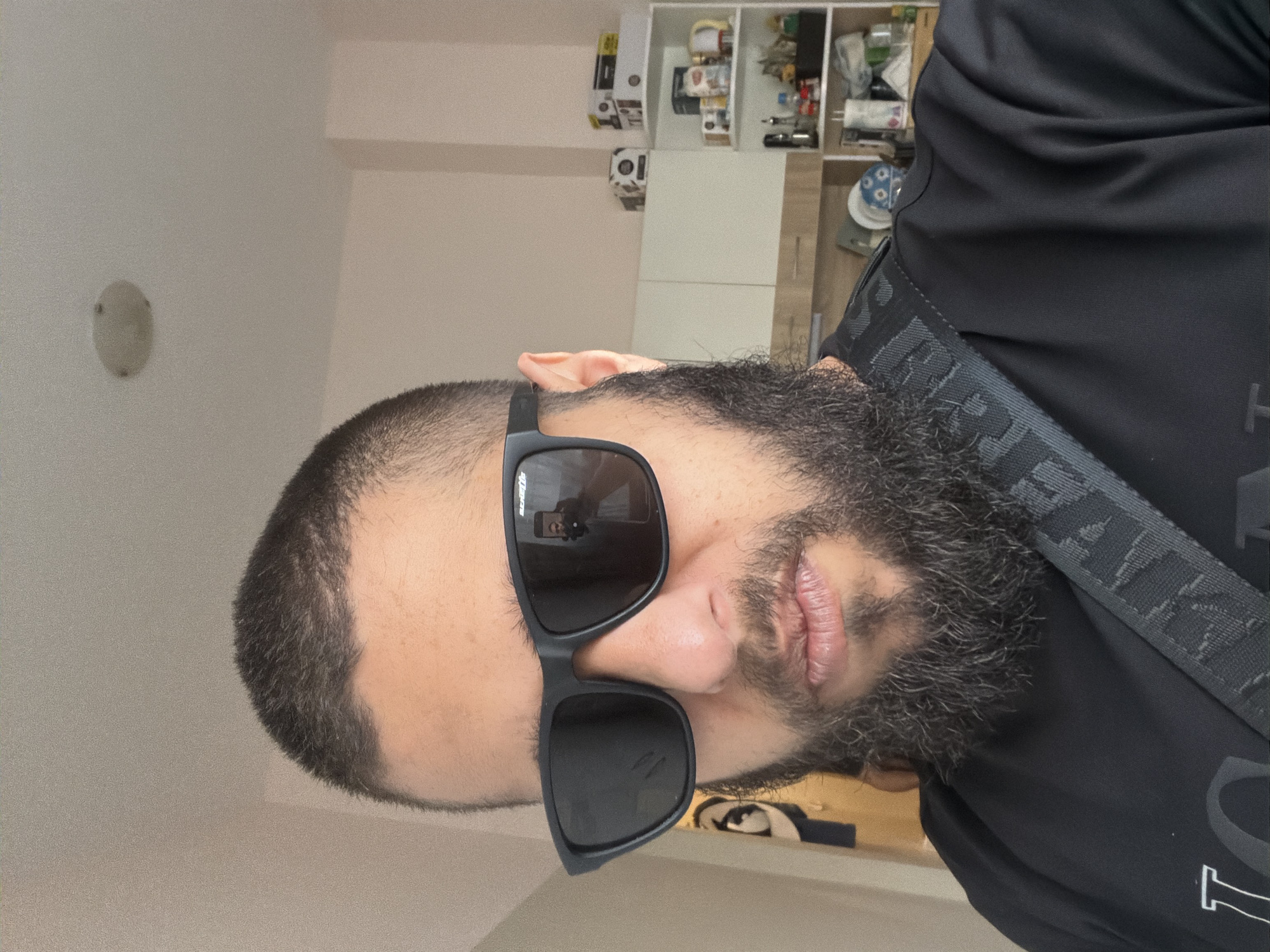 A medium shot of a man wearing sunglasses and a black shirt. The man has short hair and a full beard. He is standing indoors. In the background, there are cabinets. The location is Unknown, Unknown.