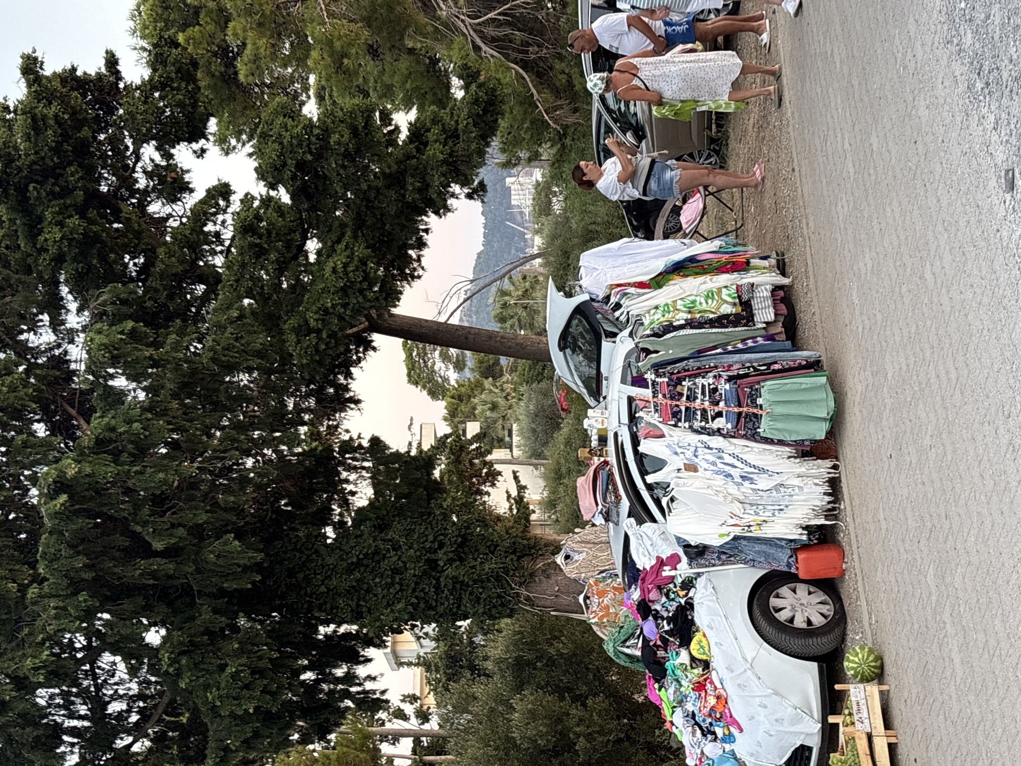 The image depicts an outdoor market scene in the city of Bar, Montenegro, with clothing and various items displayed on and around a white car. The car's trunk is open, and clothing is draped over it and piled on the hood. Racks of clothing are set up next to the car. Several people are present, appearing to be browsing or selling. In the foreground, a wooden crate contains grapes, and a watermelon is on the ground. The background shows trees, buildings, and a hilly landscape. The ground is paved with grey material. The overall impression is that of a casual street market, possibly selling used or artisanal goods.