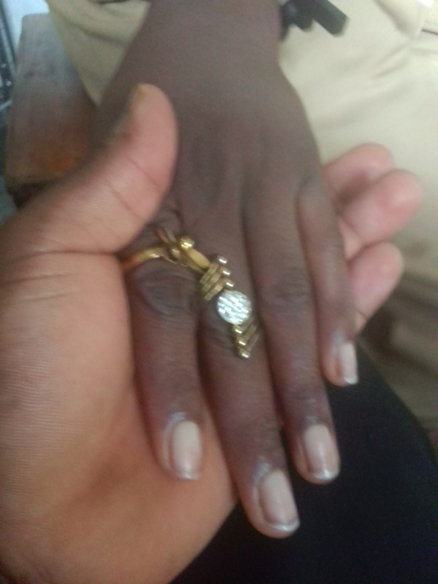 This is a close-up, eye-level shot of two hands interacting. One hand, with darker skin, is wearing a gold ring with a clear stone. The other hand, with lighter skin, is holding the first hand. The focus is on the hands and the ring. The background is blurred, showing hints of clothing and furniture. The lighting suggests an indoor setting, possibly during the daytime. The image appears to be a candid shot, capturing a moment of connection or affection between two people. There is no visible text in the image. The location is Howayi, Nigeria.