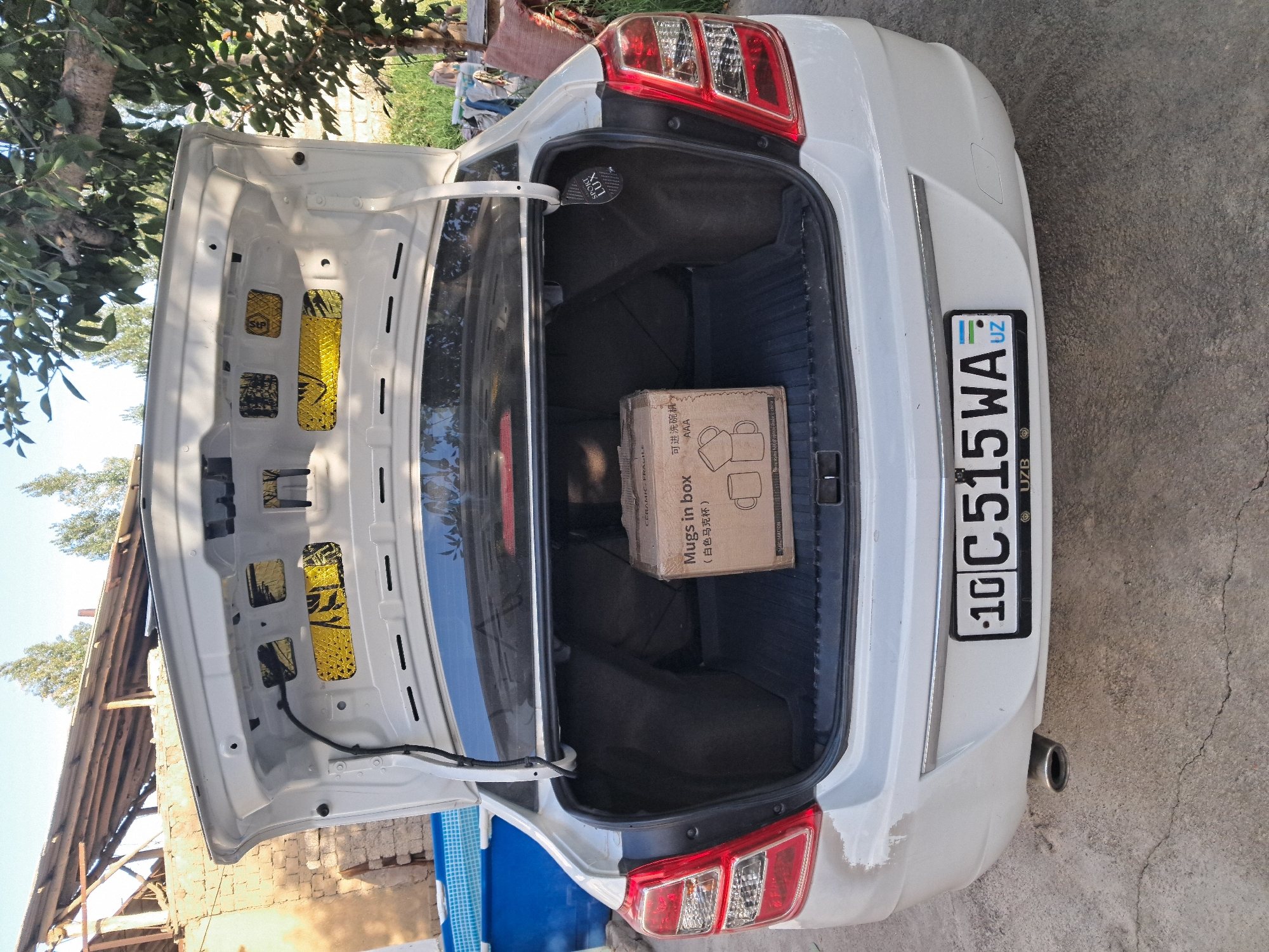 The image shows the open trunk of a white sedan parked on a concrete surface. Inside the trunk, a brown cardboard box labeled "Mugs in Box" is visible. The car's license plate, which reads "10C 515 WA UZ," is mounted on the rear bumper. The background reveals a partially constructed building with a wooden roof and some trees. The location is identified as Bagish, Uzbekistan. The scene appears to be outdoors on a sunny day.