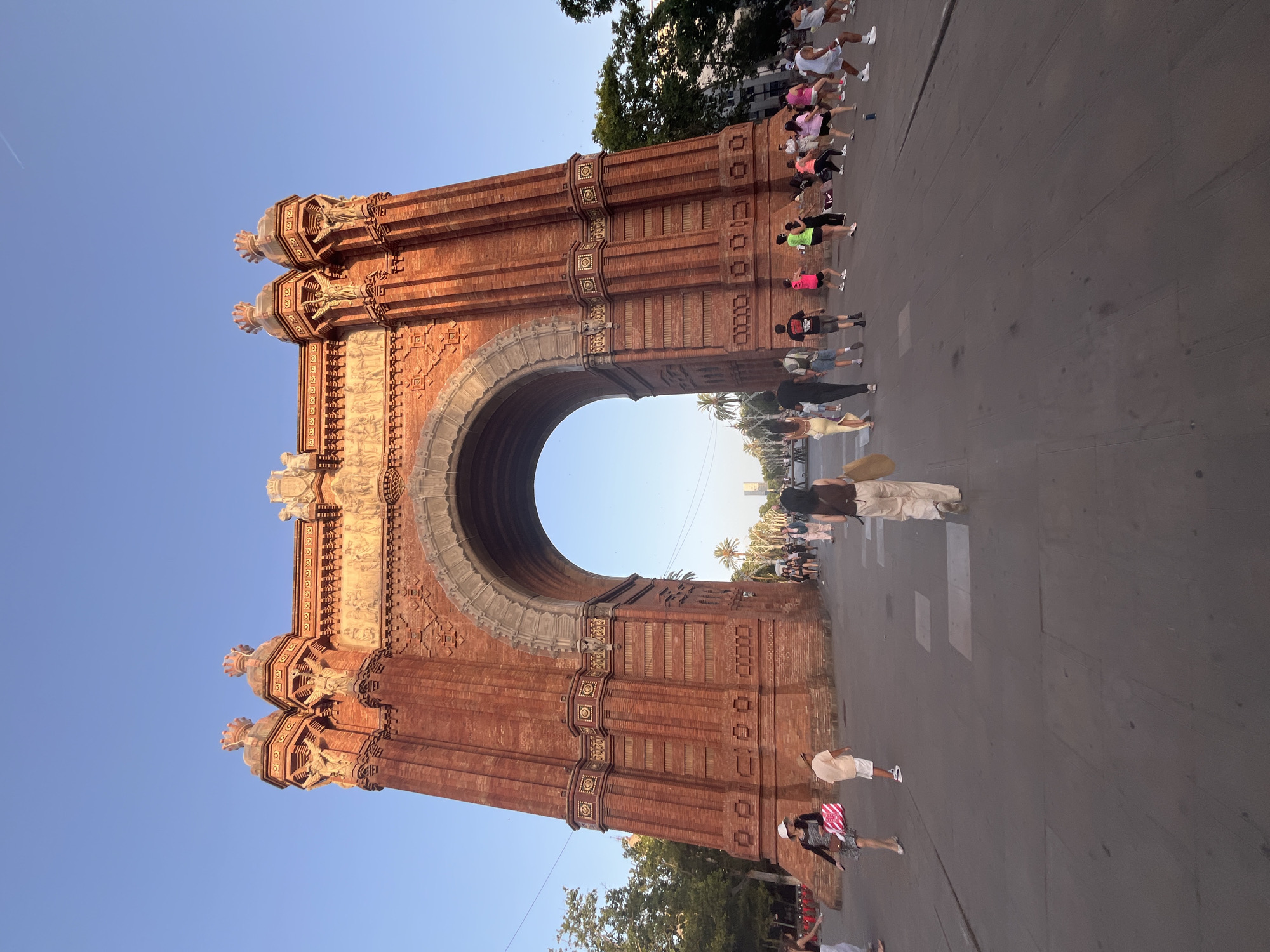 An exterior, eye-level, full shot captures the Arc de Triomf in Barcelona, Spain. The triumphal arch, constructed from red brick, dominates the center of the frame. The arch is ornately decorated with sculptures and reliefs, and is flanked by two towers. In front of the arch, a wide, paved pathway is populated by numerous people. Some individuals are walking, while others appear to be engaged in a group exercise or dance class, with several people in brightly colored athletic wear. Trees and palm trees line the pathway in the background, and a clear blue sky stretches above. The lighting suggests it is late afternoon or early evening, with a warm glow on the brickwork.