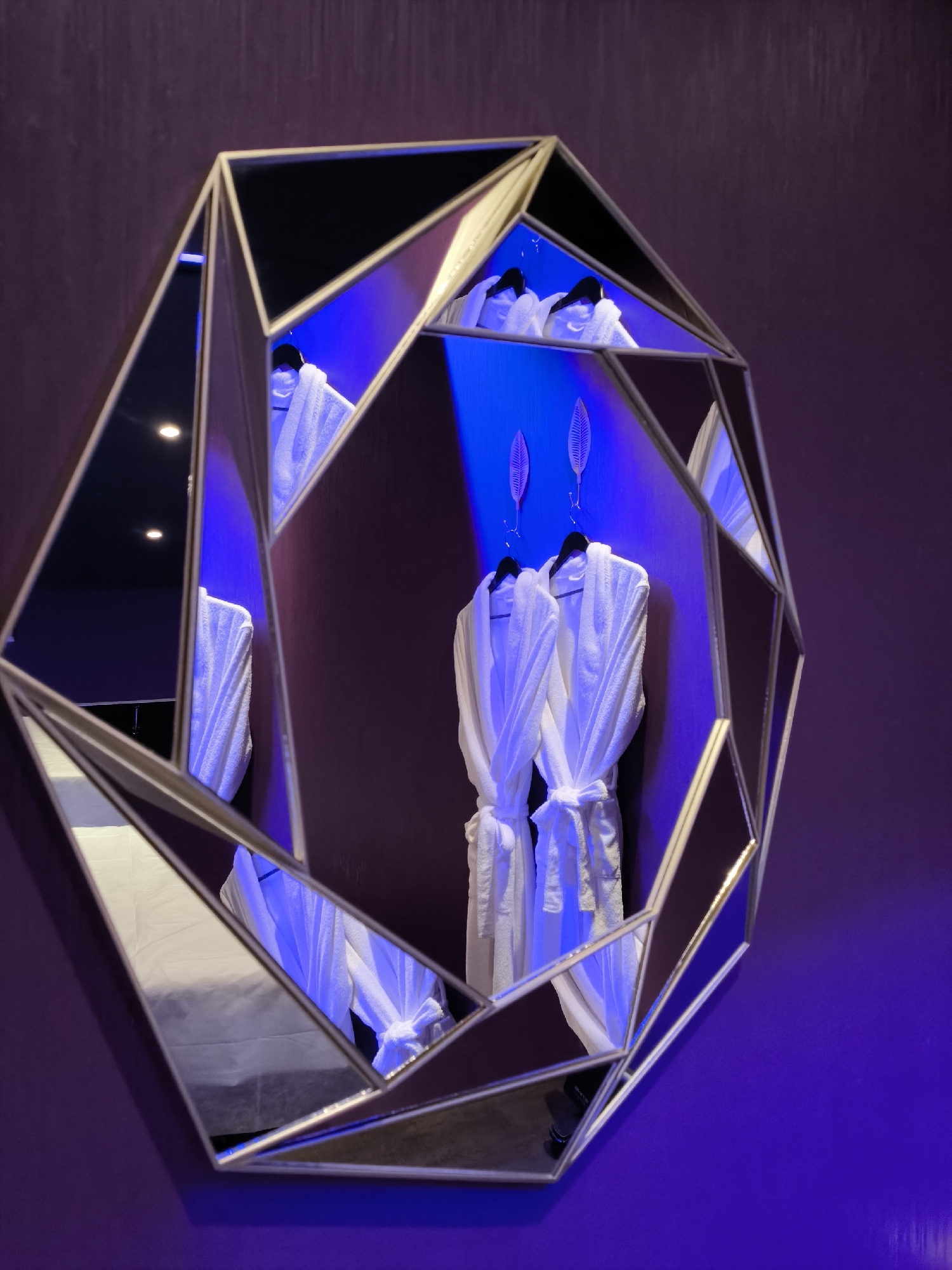 The image depicts a geometrically shaped mirror hanging on a dark purple wall, likely indoors. The mirror reflects a scene with two white bathrobes hanging on hangers, illuminated by blue lighting. There are decorative elements resembling feathers on the wall behind the bathrobes. No people are visible in the mirror or the main scene. Based on the styling and atmosphere, the location may be a high-end hotel or spa. It's challenging to confirm the location as Moscow, Russia, based solely on the image.