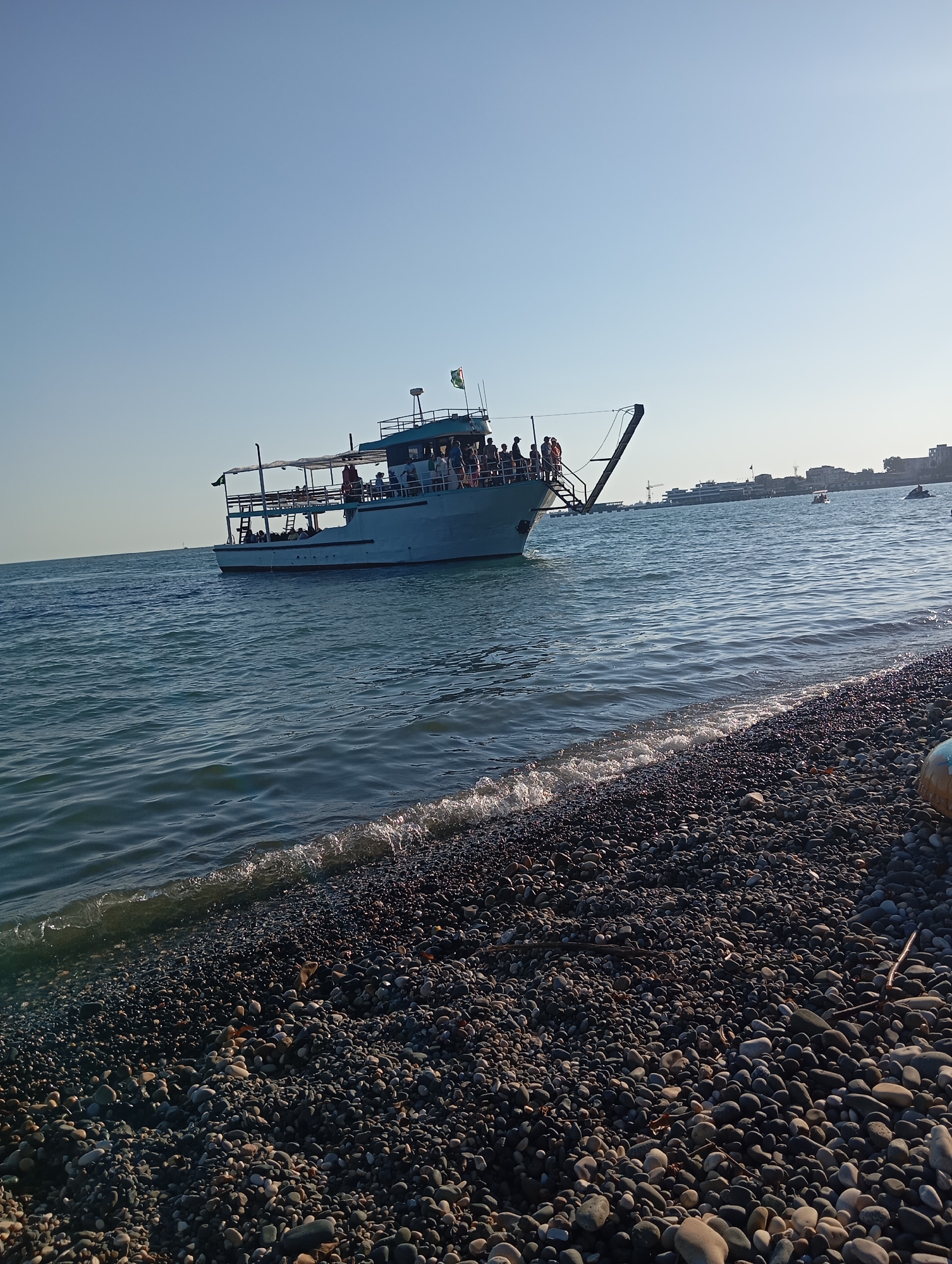 A wide-angle, eye-level shot captures a view of a large, light-colored passenger boat in the Black Sea, near the shore of Sokhumi, Abkhazia. The boat, with multiple decks and a canopy, is filled with approximately 20-30 people standing and sitting. A ramp extends from the bow of the boat, suggesting it is near or docked. The water is choppy with small waves lapping against a densely pebbled beach in the foreground. In the background, a hazy cityscape with buildings is visible on the horizon. The sky is clear and blue.
