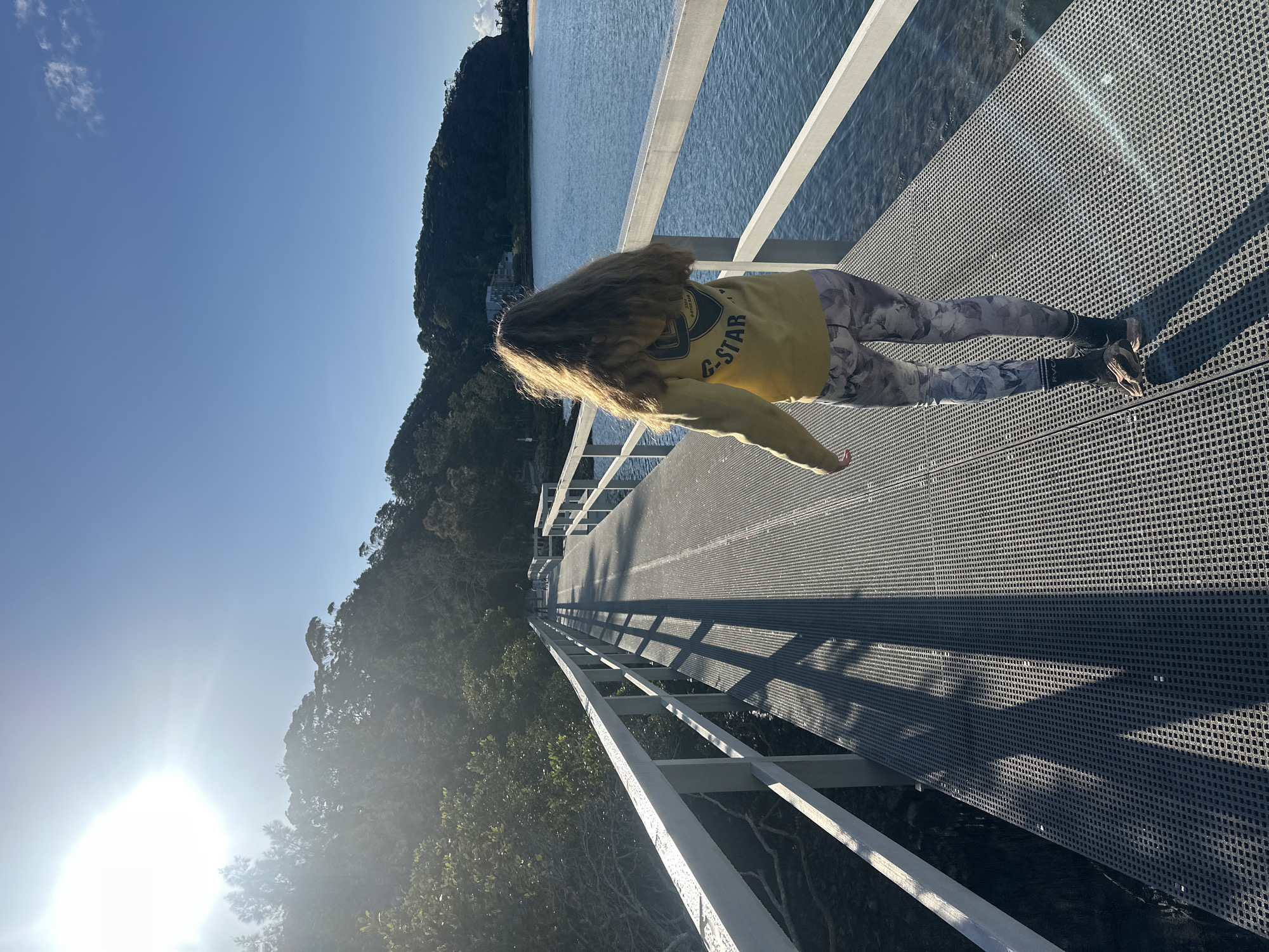 A young woman with long, blonde hair walks away from the camera on a metal boardwalk. She is wearing a yellow hooded sweatshirt with a logo on the back and patterned leggings. The boardwalk extends into the distance over a body of water, bordered by trees and hills. The sun is shining brightly in the upper left corner of the image, casting long shadows across the boardwalk. In the distance, there is a small cluster of buildings on the shore. The scene appears to be a scenic, natural setting, possibly a park or a coastal area. The location is identified as Nambucca Heads, Australia.