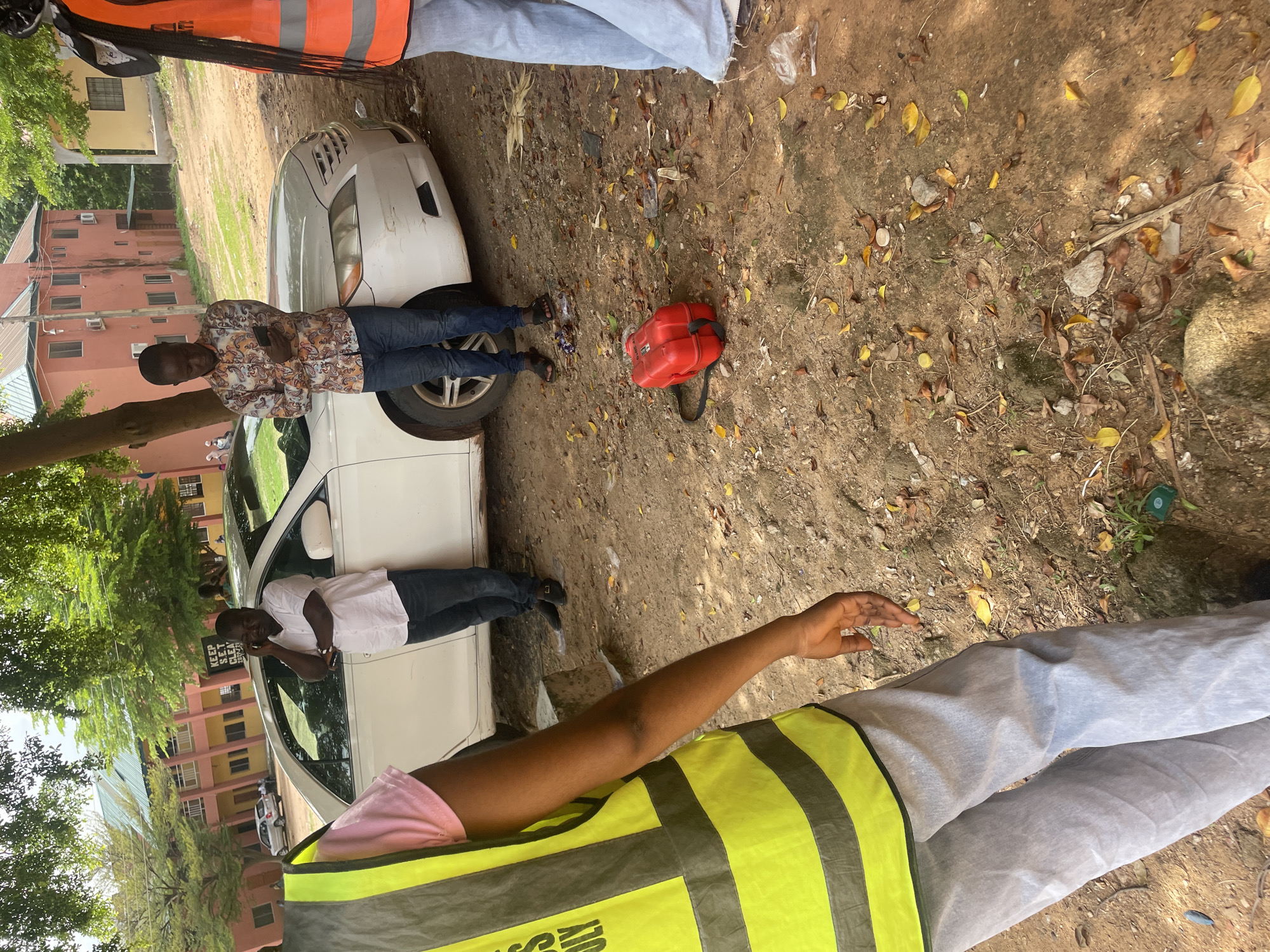 In Lokoja, Nigeria, three individuals are observed near a white sedan. One person, wearing a pink shirt and a high-visibility vest, stands facing away from the camera, their lower body and arm visible. To their right, a man in a white short-sleeved shirt and dark pants is on his phone, leaning against the car. Another man, dressed in a patterned long-sleeved shirt and blue jeans, stands near the front of the car, also looking at his phone. A red fuel canister rests on the dirt ground between the car and the man in the patterned shirt. The background reveals buildings and trees, consistent with an outdoor urban or semi-urban environment. The ground is covered with dirt, scattered leaves, and debris.