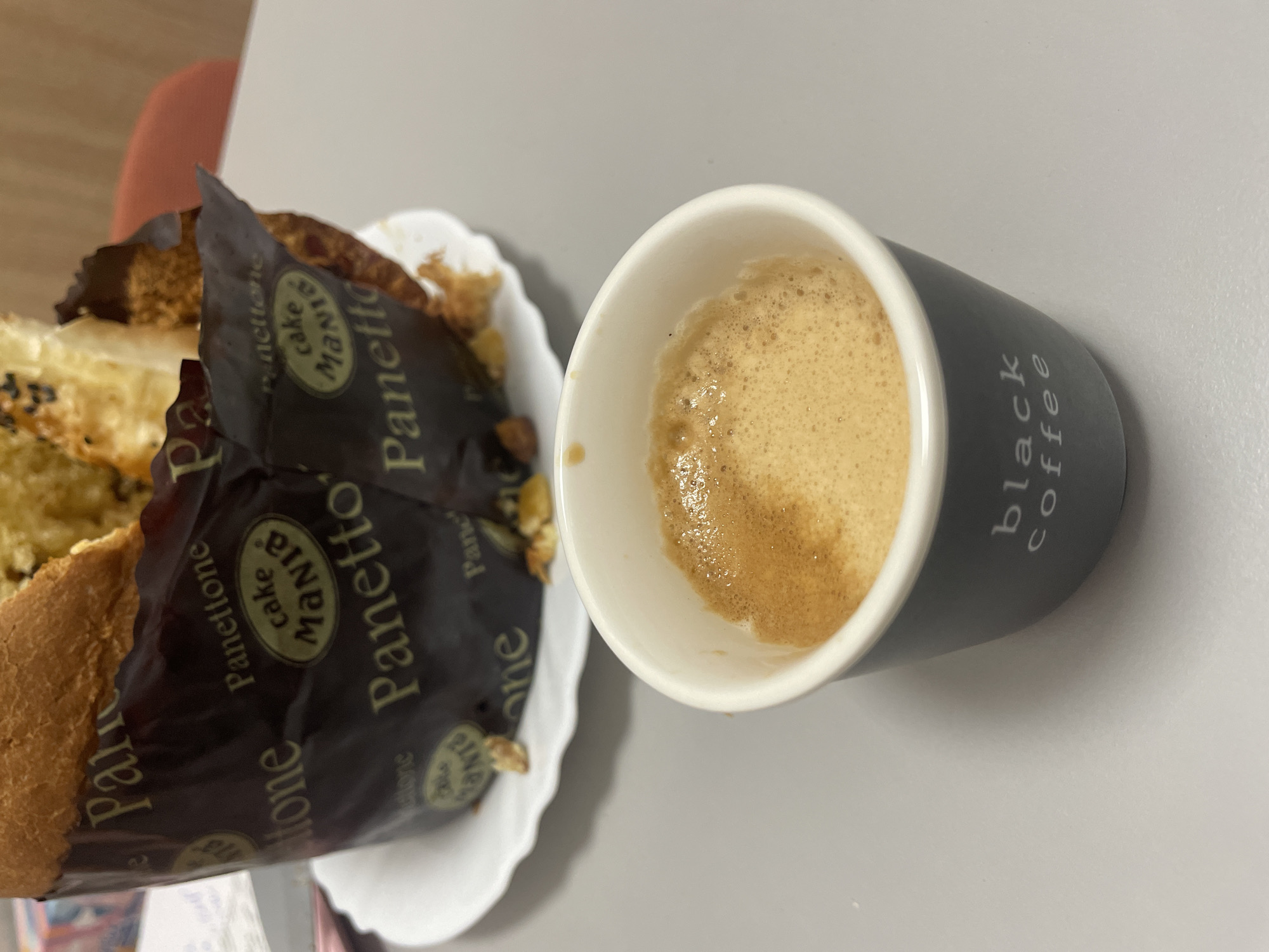 The image shows a cup of coffee and a slice of panettone on a white surface. The cup is grey and white and labeled "black coffee". The panettone slice is in its paper wrapper, which features the words "Panettone", "Mania cake", and "Panettone" again, along with other text. The picture could be taken anywhere since there is no visible context for its exact location, specifically in Sofia, Bulgaria.
