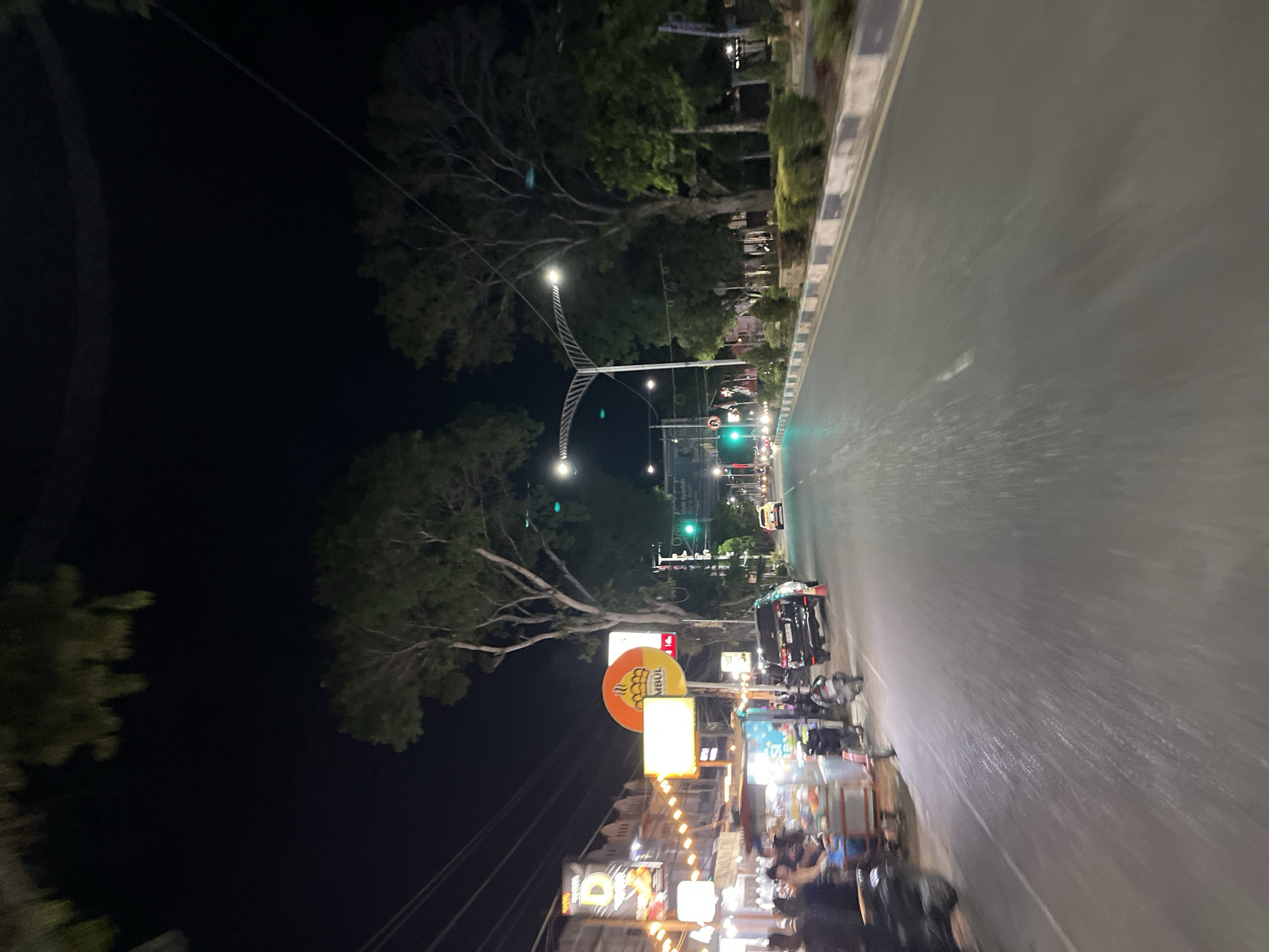 This media file captures a nighttime scene along a road in Langsa, Indonesia. The image is oriented vertically, with the road occupying the right side and a dark, tree-lined area on the left.
The road appears to be wet, reflecting the streetlights and the headlights of vehicles, suggesting it might have rained recently or is damp. Several cars and motorcycles are visible on the road, with some moving and others parked. Traffic lights are illuminated, with a green light prominently displayed in the mid-ground.
Along the left side of the road, there are numerous street vendors and food stalls, illuminated by string lights and various signs. People are gathered around these stalls, indicating a bustling night market or street food area. One notable sign features an orange circular logo with what appears to be an image of flames or a sun, and text that is partially visible and reads "MBUL". Another sign in the foreground has a large, stylized letter "D".
The presence of lush trees suggests a possibly tropical or subtropical climate. The overall atmosphere is lively and active, typical of a commercial street at night in a Southeast Asian city. The time of day is clearly night, indicated by the darkness of the sky and the reliance on artificial lighting.