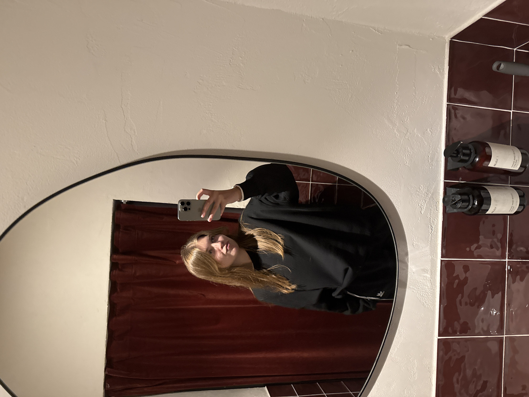 The image shows a reflection in a bathroom mirror. A young woman is taking a selfie with her phone. She is wearing a dark hoodie. The background shows a maroon-tiled wall with mounted soap dispensers. There is a curtain in the background of the reflection. The environment appears to be a bathroom, possibly in Belgrade, Serbia.