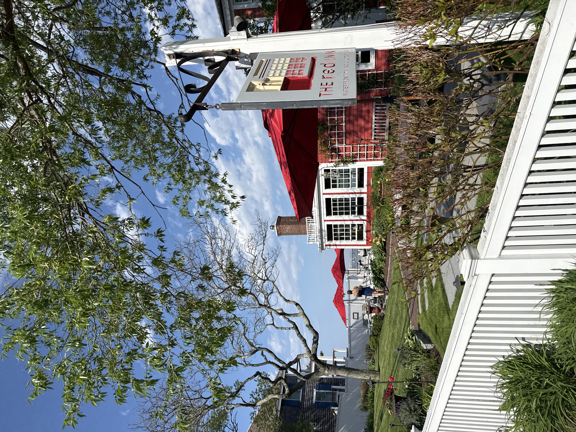 The image depicts the Red Inn, a waterfront restaurant and inn located in Provincetown, Massachusetts, United States. The building is painted red with white trim, and the sign reads "The Red Inn, Provincetown, MA 026579". Red awnings cover some of the outdoor dining areas. A white picket fence borders the property. In the background, a person is seen walking near the outdoor seating area under red umbrellas. Trees with green leaves and bare branches frame the left side of the picture, reaching towards a partly cloudy, blue sky. The environment suggests a pleasant, summer day.