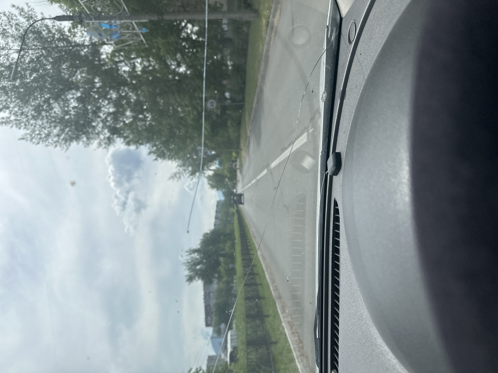 The image shows a view through a car windshield. The windshield has several cracks and is dirty. The car is on a paved road with white lane markings. On the left side of the road, there is a metal fence and then some buildings in the distance. On the right side of the road, there are trees and a lamppost with a speed limit sign that appears to indicate 60. The sky is overcast with clouds. The dashboard of the car is visible in the foreground. The location appears to be Sayanogorsk, Russia, based on the context provided.