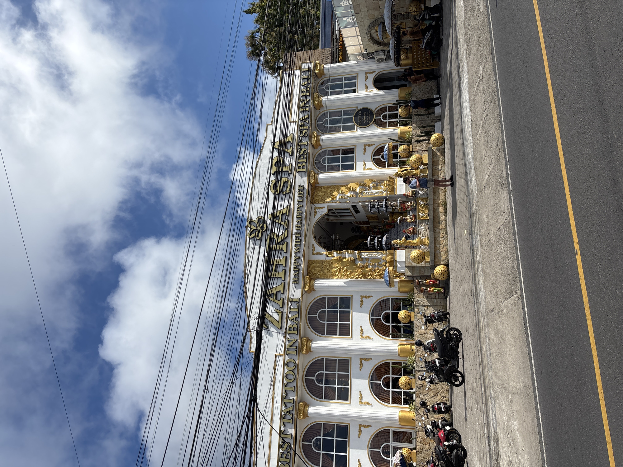 The image captures a view of the exterior of a building in Ungasan, Indonesia. The building, identified as a tattoo studio and spa, is decorated with ornate gold detailing. A number of motorcycles are parked on the pavement in front of the building. Above the building are utility cables against a backdrop of a partly cloudy blue sky. A road runs alongside the pavement with a yellow centerline. Several people are standing or walking around the building.
