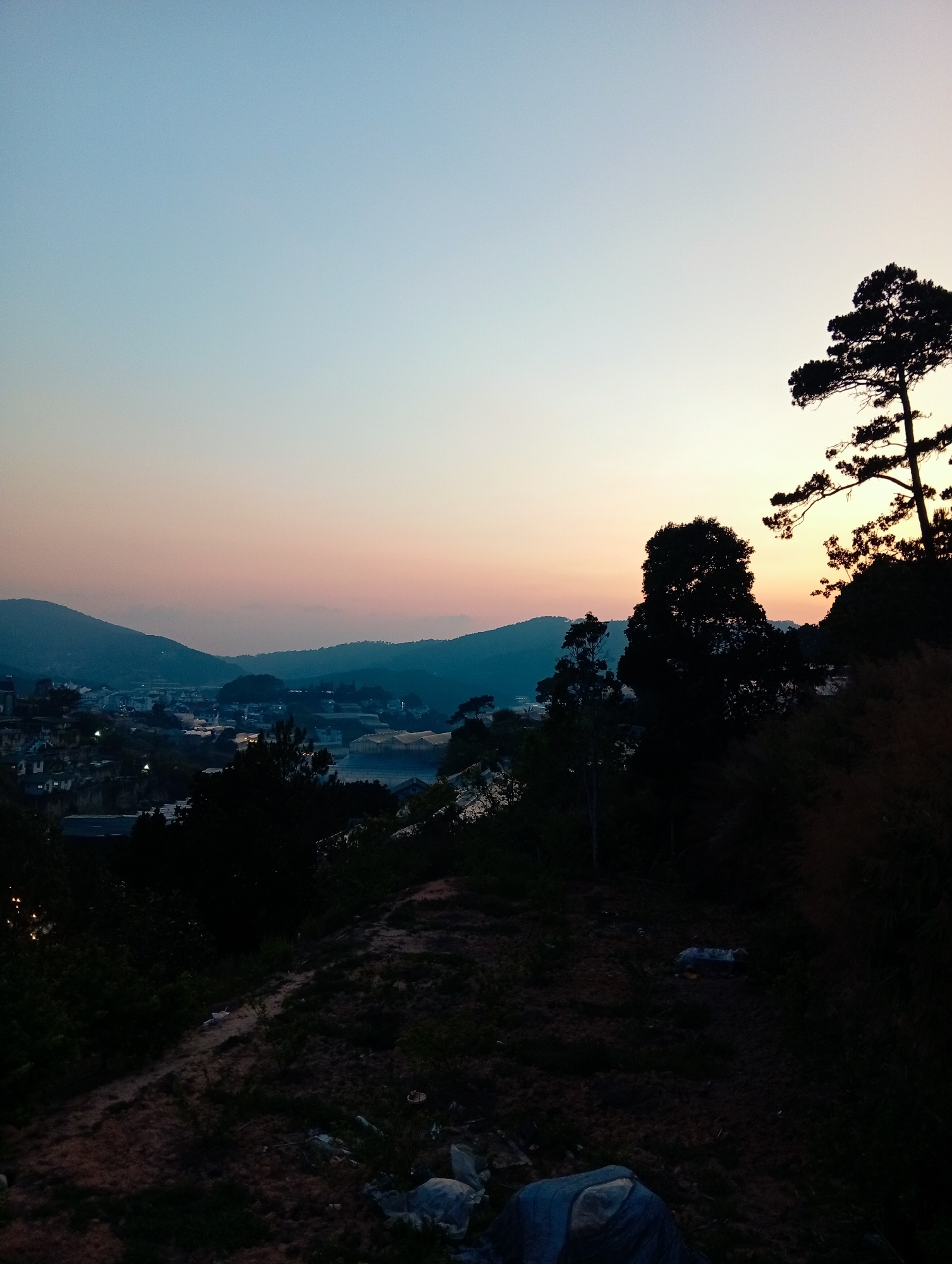 The image captures a landscape scene at dusk in Da Lat, Vietnam. In the foreground, there's a dark, overgrown slope with vegetation and scattered debris, including what appears to be trash. The midground features silhouettes of trees against the colorful sky, with a view of the city nestled amidst rolling hills and mountains. Lights from the city faintly illuminate the landscape. The sky transitions from a soft blue at the top to a gradient of orange and pink near the horizon.