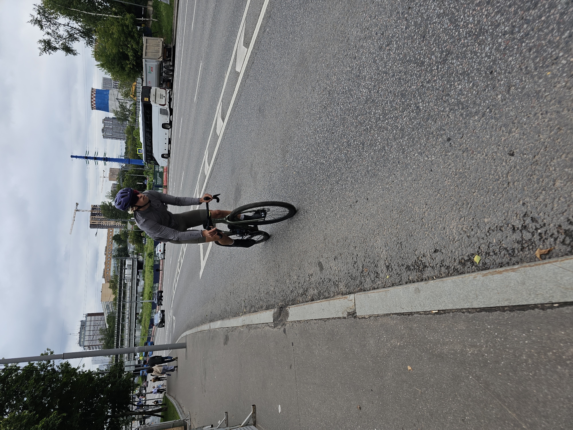 A man wearing a helmet and cycling attire is riding a bicycle on an asphalt road in Moscow, Russia. He is wearing a grey long-sleeved shirt and dark shorts, and a purple helmet. The man is leaning forward over his handlebars. The road has white lane markings. To the left of the road is a sidewalk with several pedestrians walking. In the background, there are buildings, trees, a white bus, and a semi-trailer truck. The sky is overcast.
