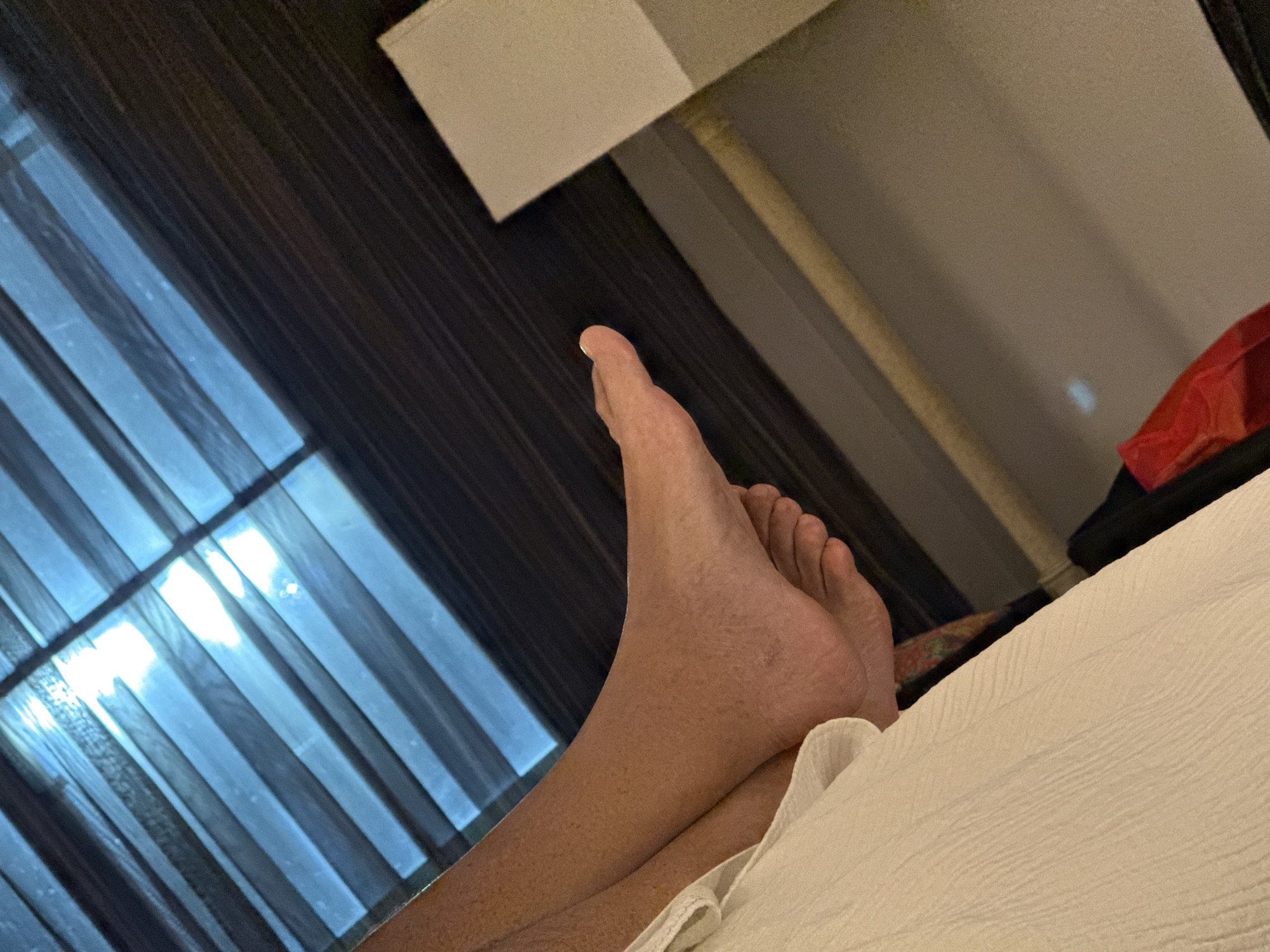 The image shows a person's bare foot resting on a white bedsheet. The foot is positioned in the lower center of the frame, with the toes pointing upwards. A second leg is seen beneath the bedsheet. The background includes a window with dark curtains, allowing light to filter through. There is also a bedside table and a glimpse of a red object. The location cannot be determined from this image, and there is no clear evidence that the image was taken in San Antonio, United States of America.