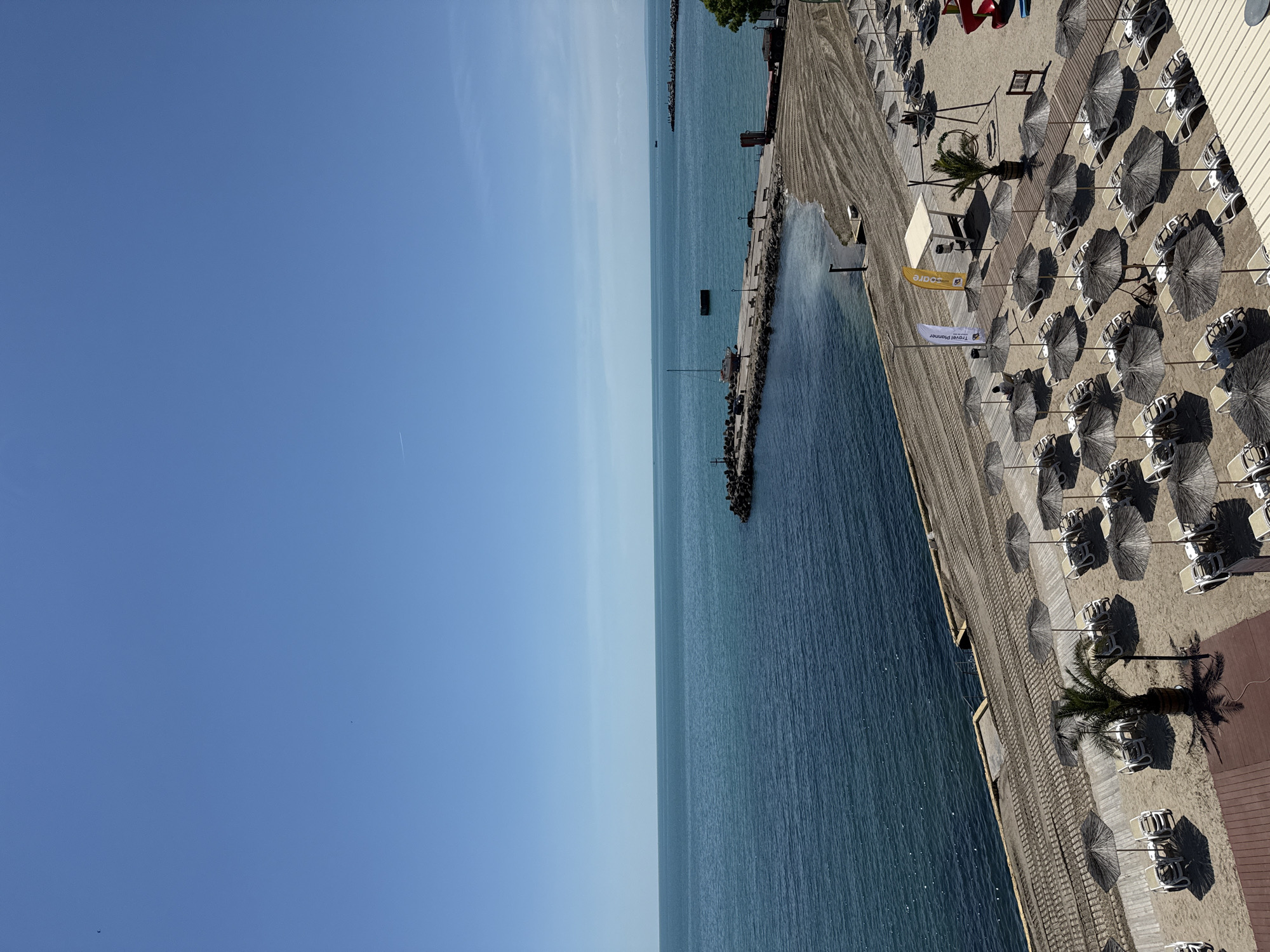 The image depicts a beach resort area in Kavarna, Bulgaria, seen from an elevated perspective, potentially from a high building. The scene is primarily focused on a stretch of coastline with a blue sea on the left, gradually transitioning to a clear, light blue sky. A pier extends into the sea, bordered by rock structures, likely serving as a breakwater. To the right of the sea, a sandy beach is arranged with rows of thatched umbrellas and sunbeds. A wooden boardwalk runs along the edge of the beach, parallel to the shoreline. Several palm trees are strategically planted amongst the umbrellas, providing shade. Flags and banners are visible near the edge of the beach, suggesting promotional or branding elements. The overall impression is a tranquil beach scene during a clear day.