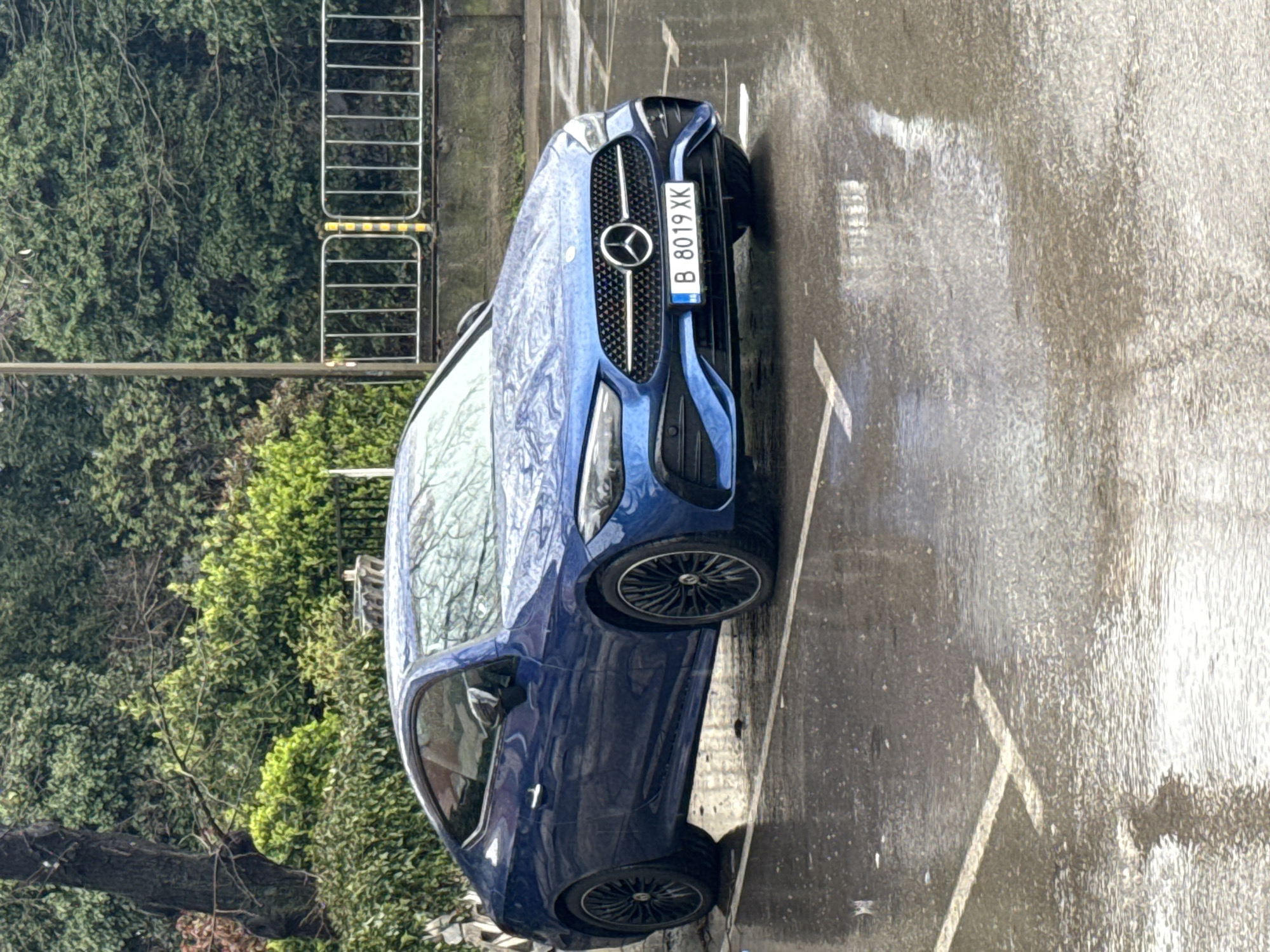 A blue Mercedes-Benz is parked in a designated parking space on a wet street, likely in an urban area. The car has a license plate that reads "B 8019 XK". The vehicle is positioned at an angle relative to the parking space lines. Lush green foliage, possibly trees or bushes, is visible in the background, along with a metal barrier. The wet pavement suggests recent rainfall, and reflections of the surroundings are visible on the car's surface. Based on the license plate, the car is registered in Bucharest, Romania, not Varna, Bulgaria. The image is shot from a slightly elevated angle.