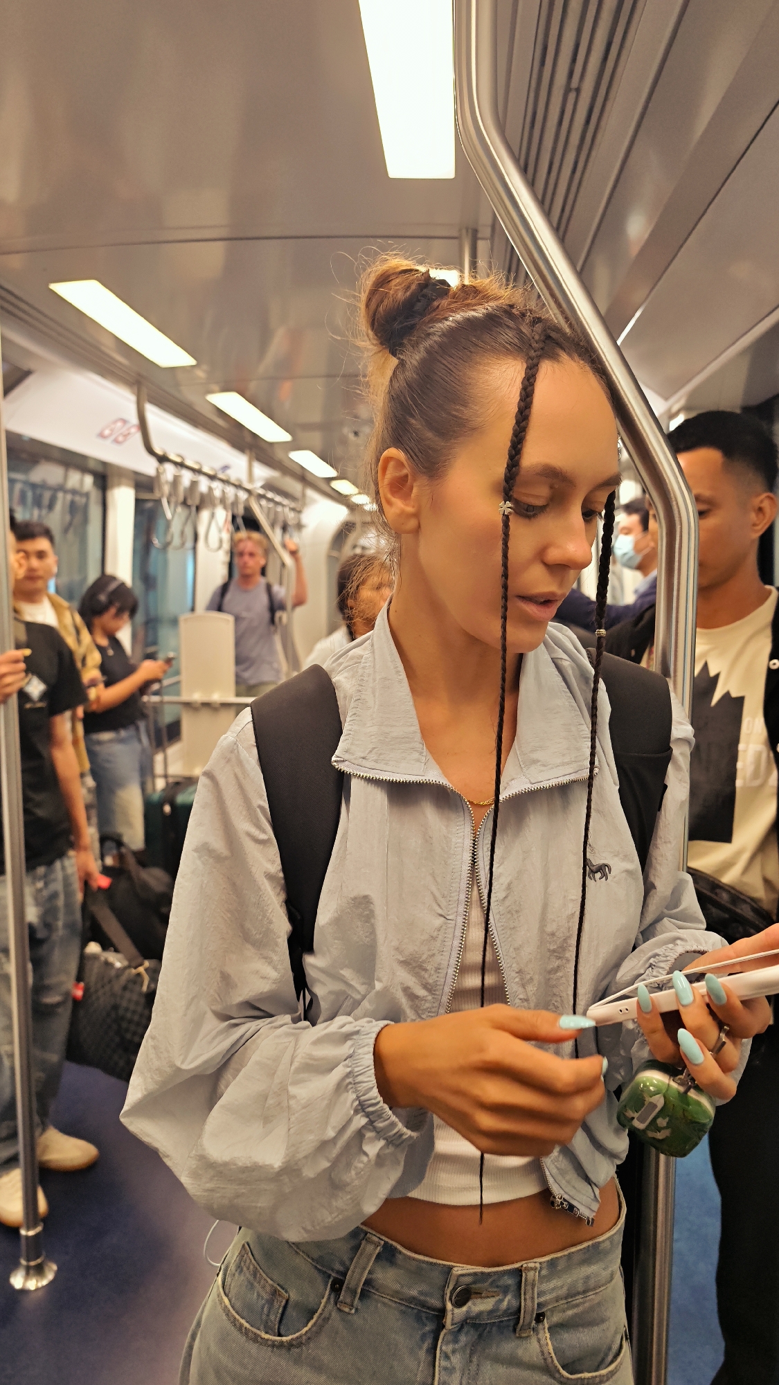 A young woman with her hair in a top knot and braids stands on a train, holding a smartphone and a green case. She is wearing a light blue zippered jacket over a white crop top and light blue jeans. Her fingernails are painted a light blue. The woman is looking down at her phone, her expression focused.
In the background, other passengers are visible on the train. A man in a white t-shirt stands to her right. To her left, a man in a yellow plaid shirt and another person with dark hair and a blue shirt are visible. The train car is brightly lit by fluorescent lights along the ceiling. Handrails are present for passengers to hold onto. Luggage can be seen near some of the other passengers. The setting appears to be a public transportation vehicle, likely a train or metro, in an indoor environment. The overall impression is of a casual journey, possibly during the day given the lighting. No specific weather conditions are discernible. No visible text is present in the image. The location context is Nong Prue Subdistrict, Thailand.