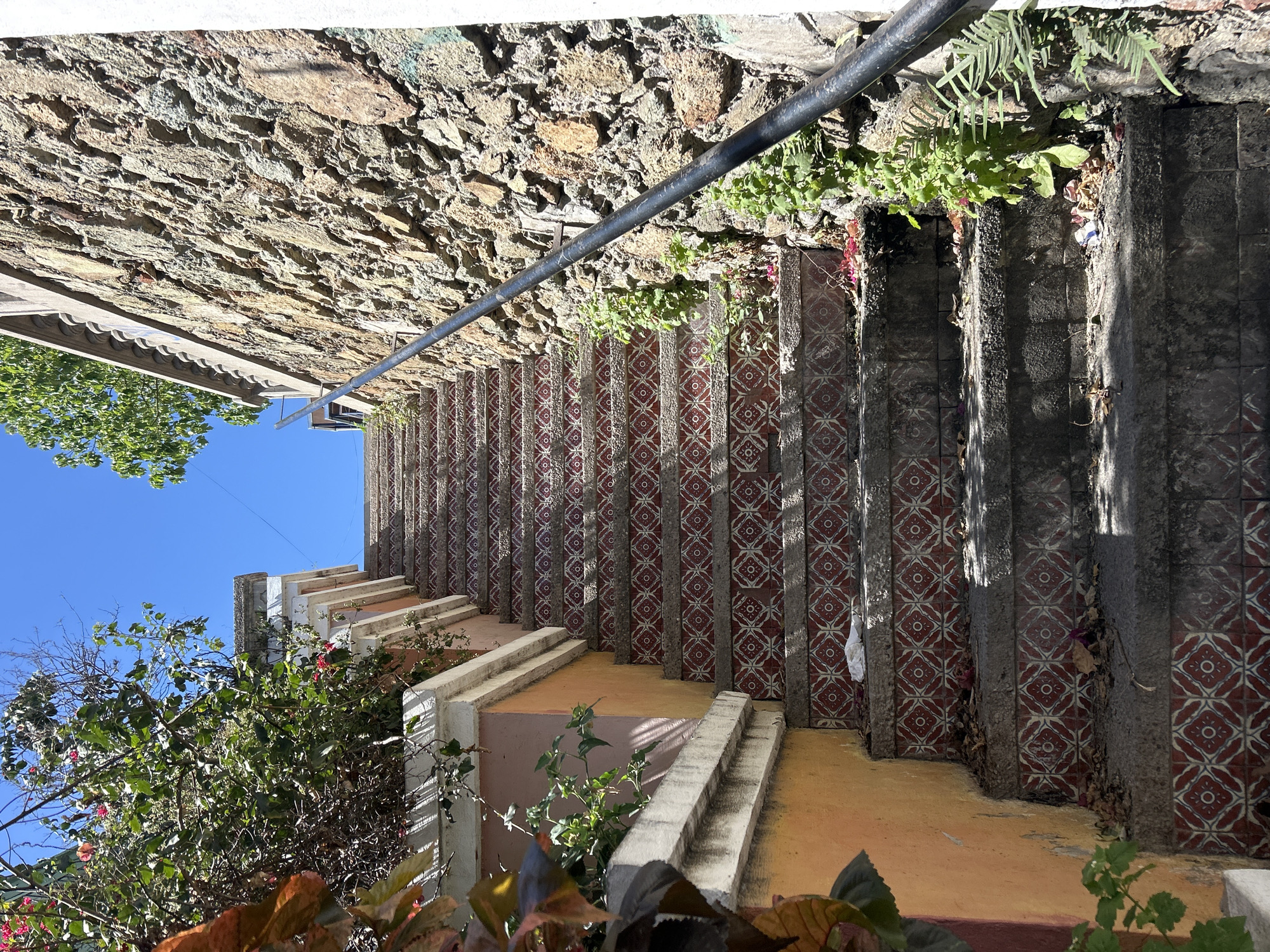 The image depicts a set of outdoor stairs in Mazatlán, Mexico. The stairs ascend vertically in the frame, constructed with patterned tiles featuring red and white geometric designs. A black metal railing runs along the upper left side of the stairs. The stairs appear to be worn and weathered, with some debris accumulated along the sides. A stone wall borders the upper left of the stairs, with greenery growing along the side. The bottom of the frame shows plants and foliage in front of the stairs. Above the plants, a clear blue sky and some greenery are visible in the background.