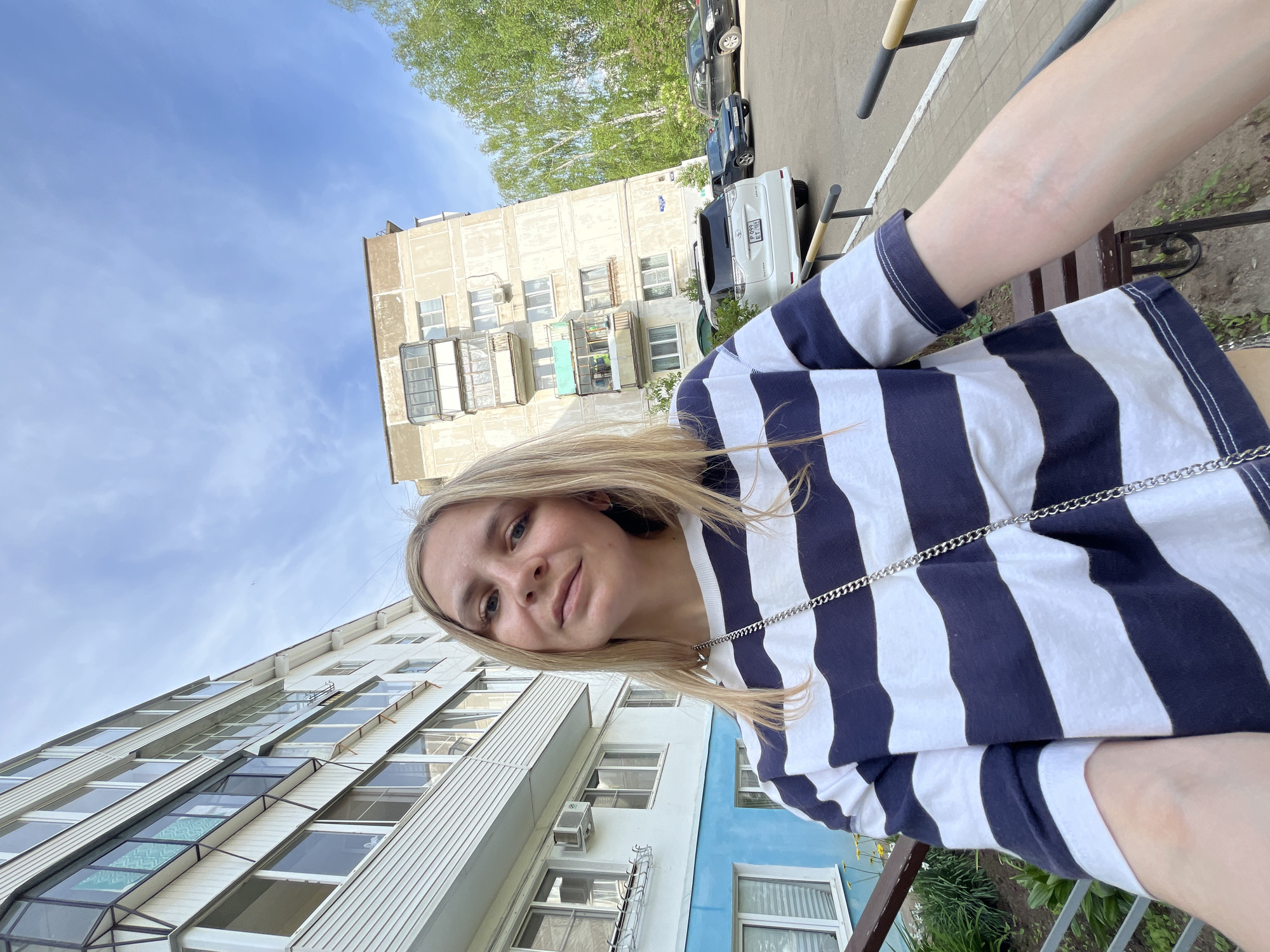 A medium-shot, rotated selfie of a young woman with blonde hair is set against a backdrop of urban architecture and a partially visible sky. The woman is wearing a navy blue and white striped t-shirt, and a silver chain is visible around her neckline. She is positioned at an angle, with her face tilted towards the camera.
Behind her, two multi-story buildings are visible. The building in the lower-left is painted white with blue accents and features numerous windows, some of which have air conditioning units installed. The building in the upper-center is a more conventional apartment block, likely made of concrete. Trees are visible behind the building in the upper-center.
In the background, a parking area with several parked cars can be seen. A bench is visible near her arm, as well as some landscaping.
Based on details such as the architectural style of the buildings and the license plates on the vehicles in the background, this image was likely taken in the city of Chaikovsky, Russia.