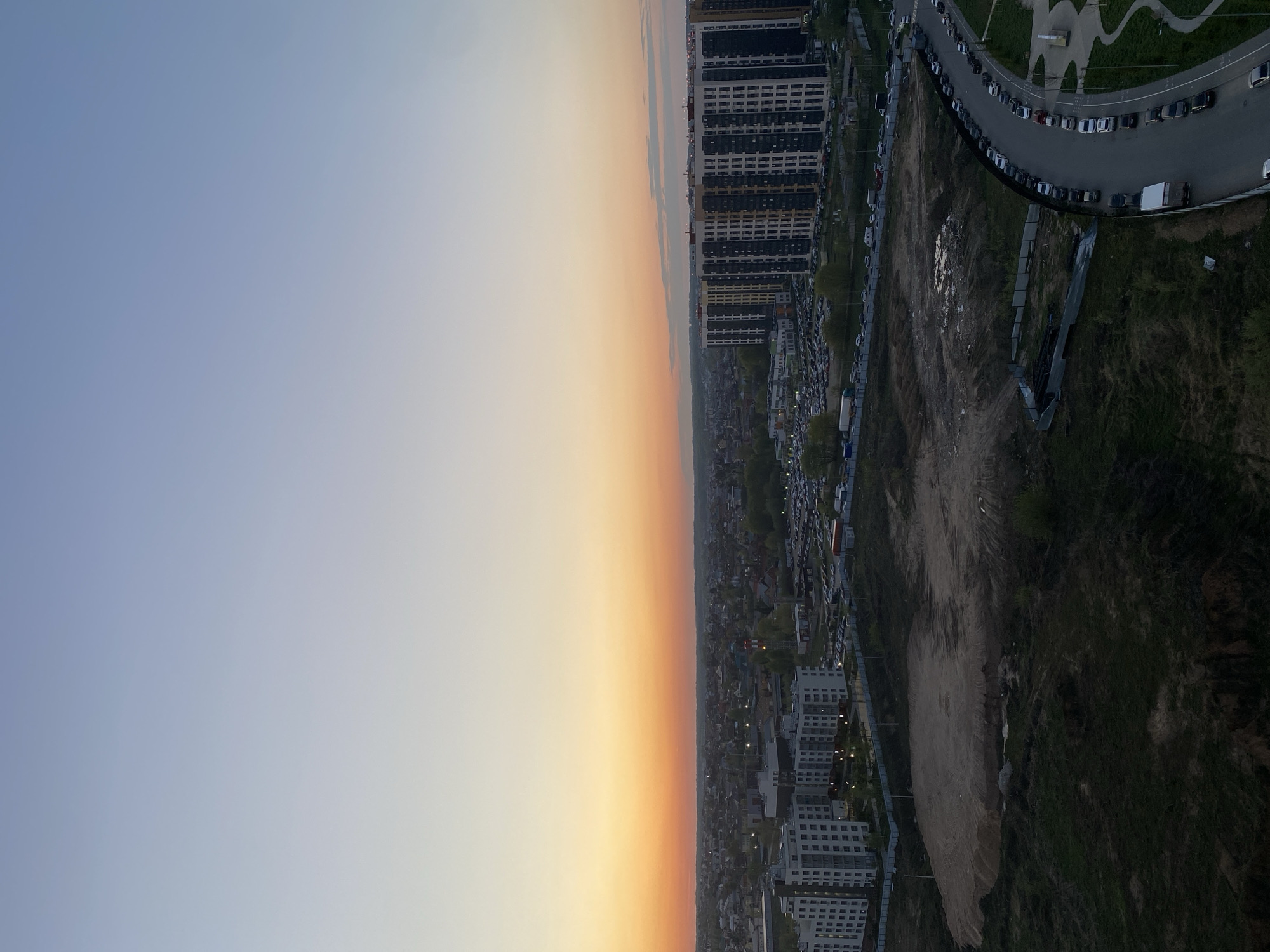 The image shows a cityscape with a clear sky gradient transitioning from light blue to orange. A mix of high-rise apartment buildings and lower residential structures lines the horizon. There appears to be a parking area along a curved street, filled with vehicles, and a vacant lot with exposed dirt and overgrown grass.
The photograph was taken in Kazan, Russia.