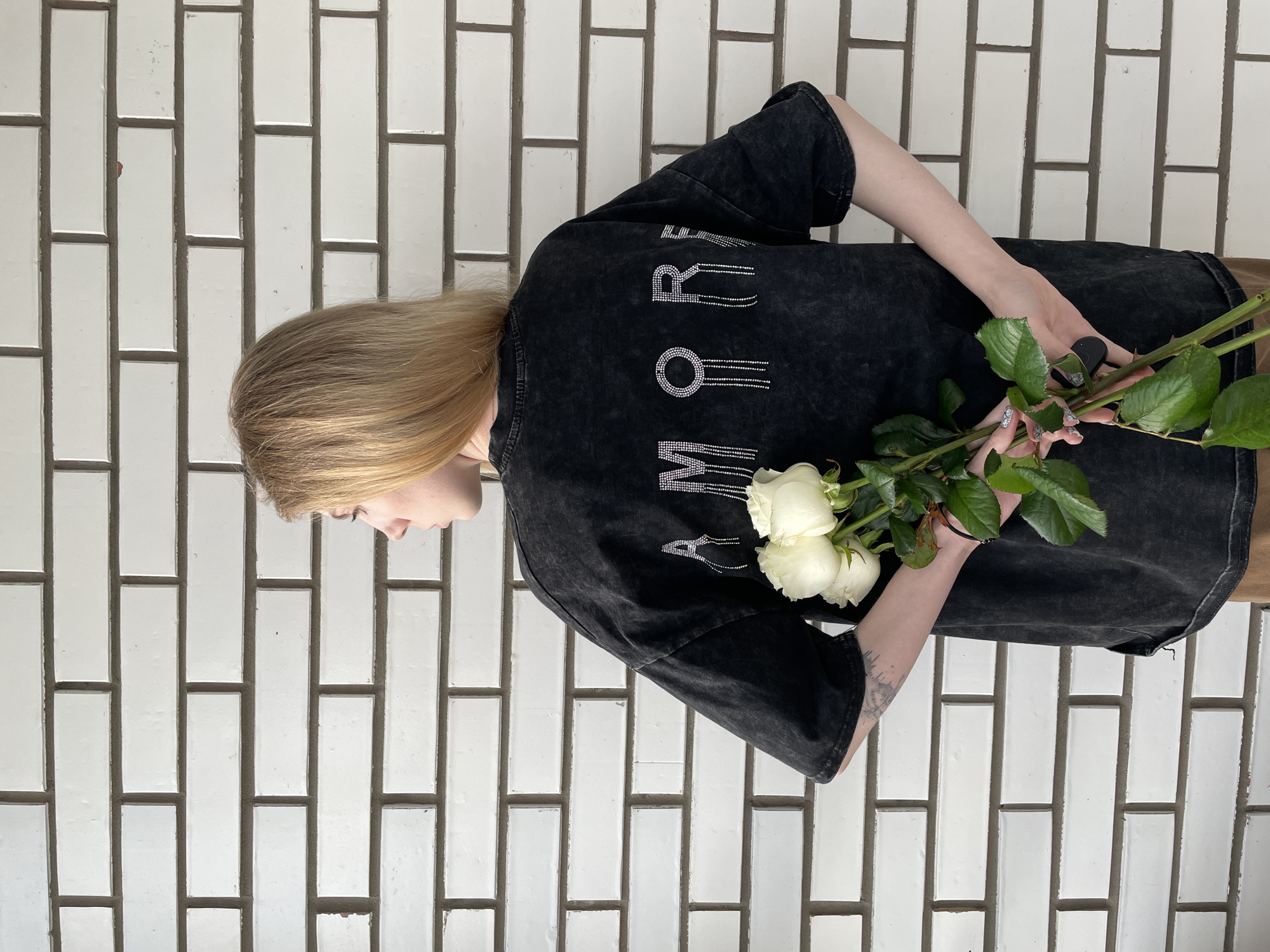 A person is holding a bouquet of white roses behind their back. The individual is wearing a black t-shirt with the word "AMORE" printed on the back in a vertical line with rhinestone accents, and has blonde hair. The backdrop is a wall composed of vertically oriented, white rectangular tiles with dark grout. The location is not discernible in the image.