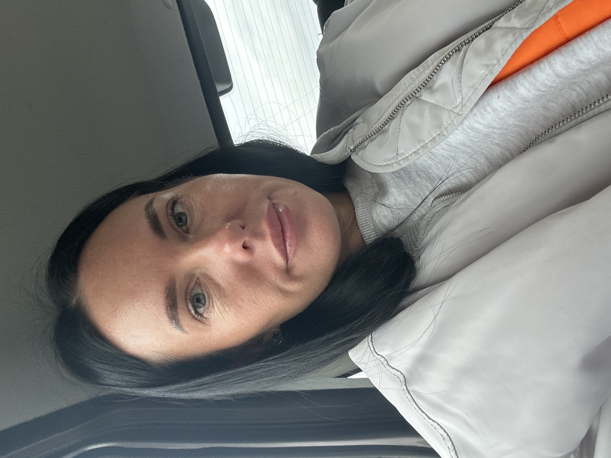 A woman with dark hair and fair skin is shown in a close-up shot. She is wearing a light gray quilted jacket over a gray sweatshirt with a glimpse of an orange layer underneath. The background suggests she is inside a car due to the presence of a car's ceiling and window frame. There's no indication of the location being Ольховка, Russia.