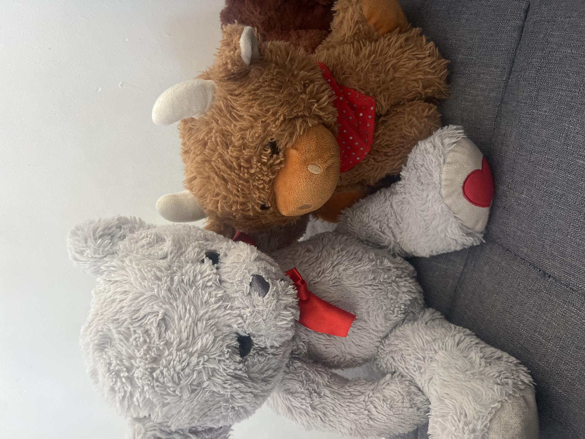 The image depicts two stuffed animals, a brown moose with a red polka-dotted bandana, and a gray teddy bear with a red ribbon, resting on a gray fabric surface. The scene is devoid of human presence and provides no discernible clues regarding its location within Gorleston-on-Sea, United Kingdom. The environment is limited to the textures and colors of the stuffed animals and the gray surface they are on. There are no actions or interactions visible, only the still life composition of the objects.