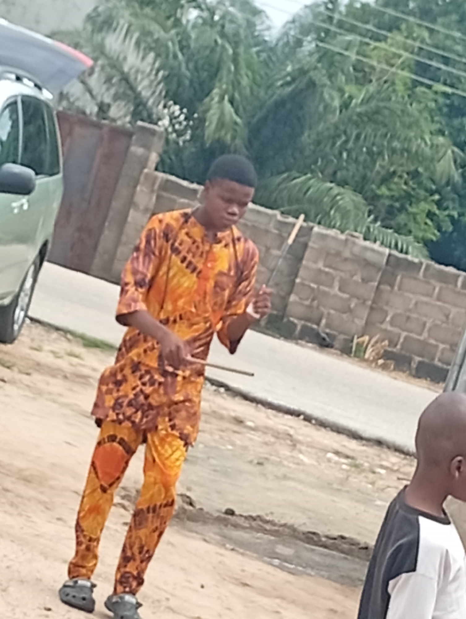 In an outdoor urban environment, a young Black male wearing a vibrant orange and brown patterned traditional African outfit and grey Crocs is captured mid-stride. He is looking down and to his right, with his right hand raised in a gesture that could be a wave or a dance move. Behind him, a light-colored SUV is parked, partially obscuring a brown wooden gate and a grey brick wall. Lush green foliage, likely palm trees, forms a dense backdrop against a lightly overcast sky. To the right of the main subject, another young Black male, with a shaved head and wearing a black and white t-shirt, is partially visible, facing away from the camera. The ground is covered in sand and dirt, suggesting a less developed or under construction area.
